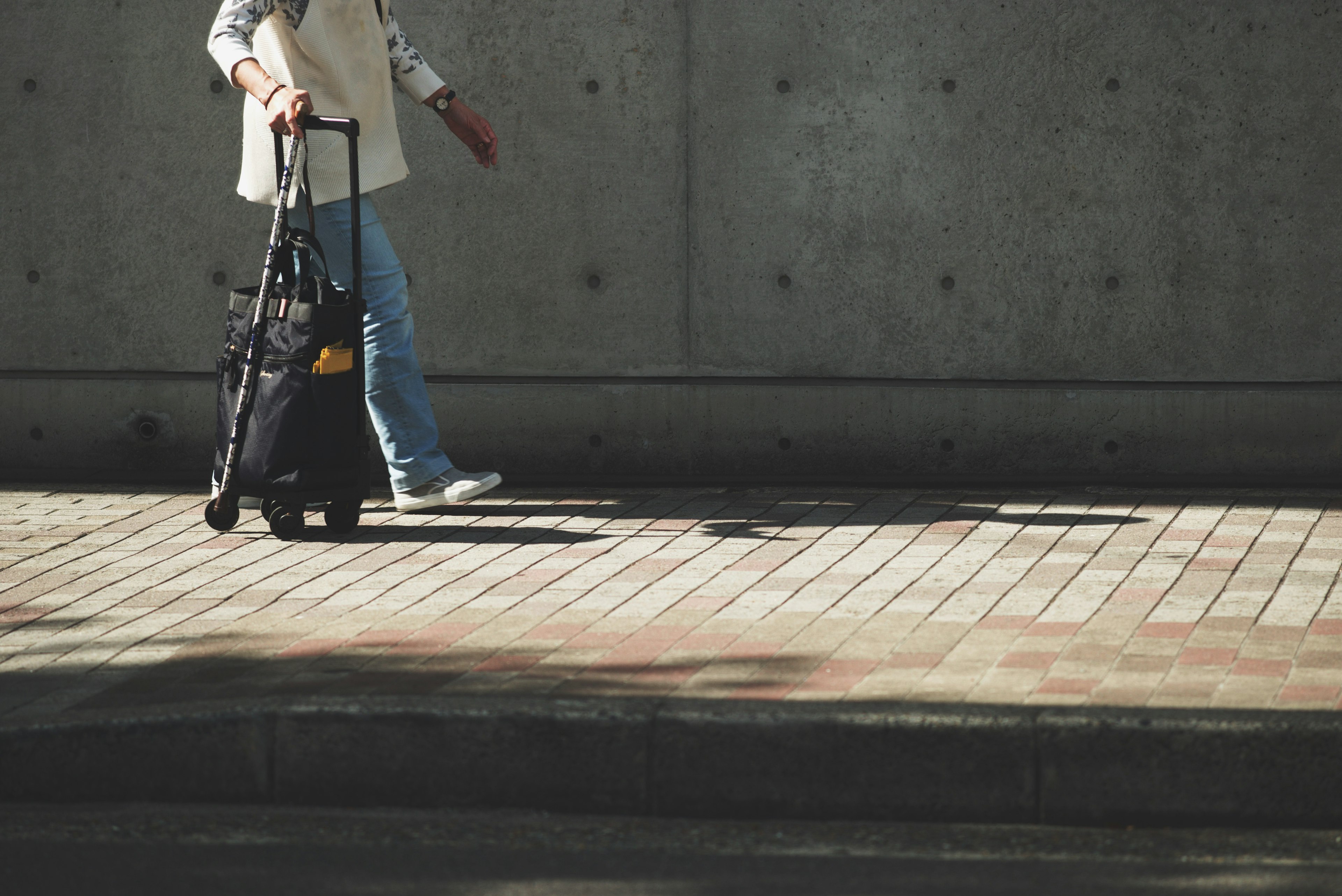 A person walking while pulling a wheeled bag against a concrete wall and paved sidewalk