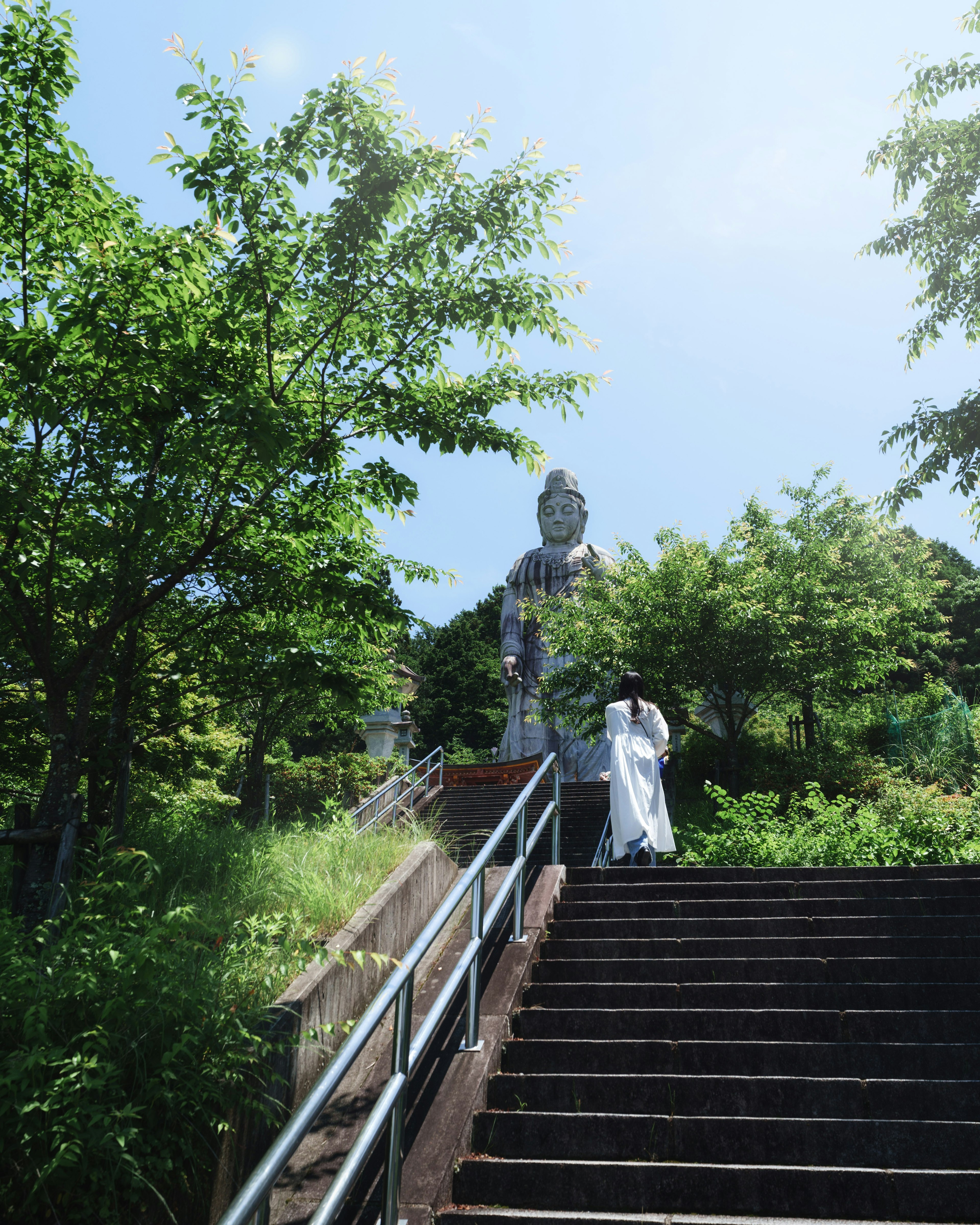 A person in white clothing ascending stone steps surrounded by greenery with a large Buddha statue in the background