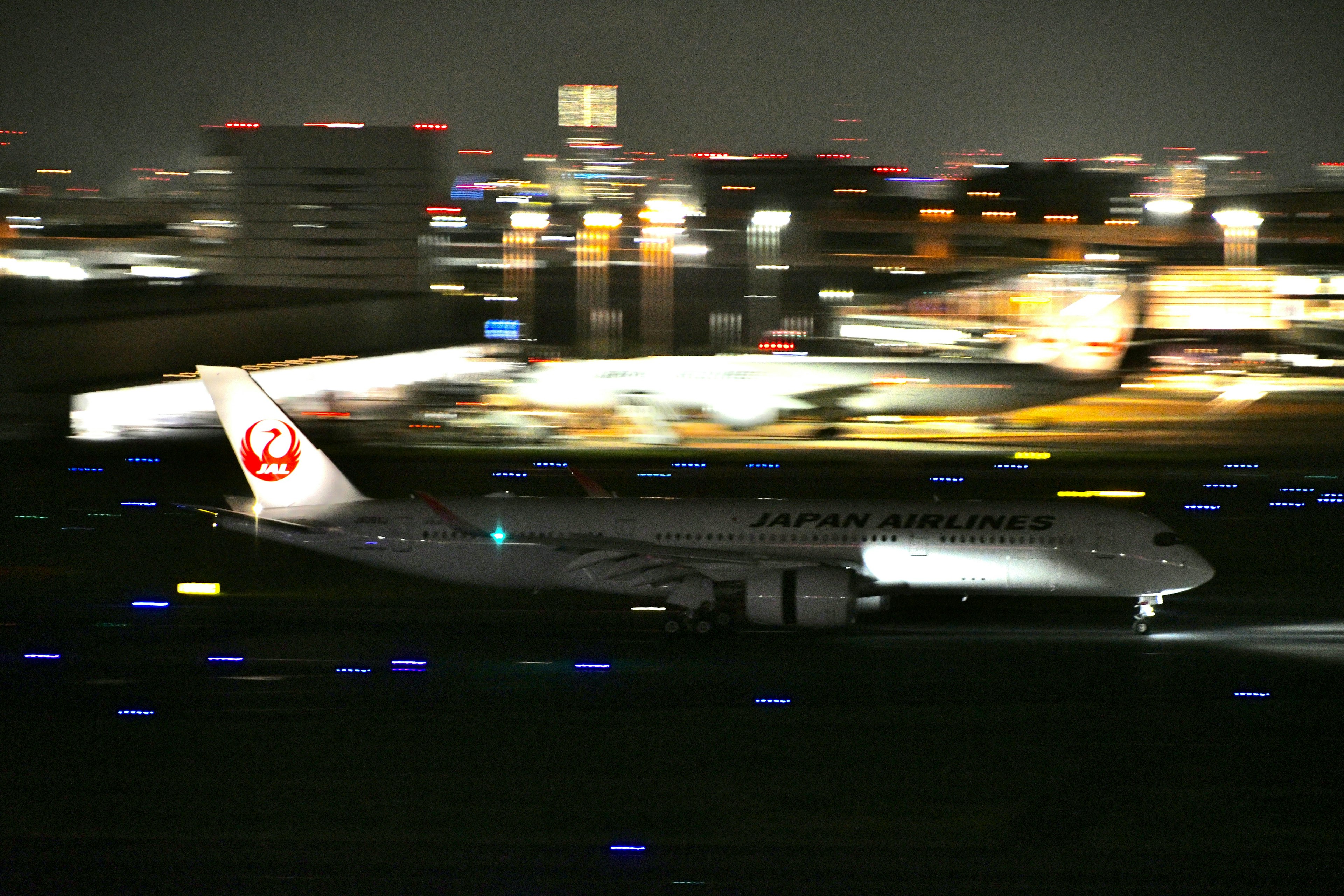 Pesawat Japan Airlines lepas landas di malam hari dengan latar belakang kota yang kabur