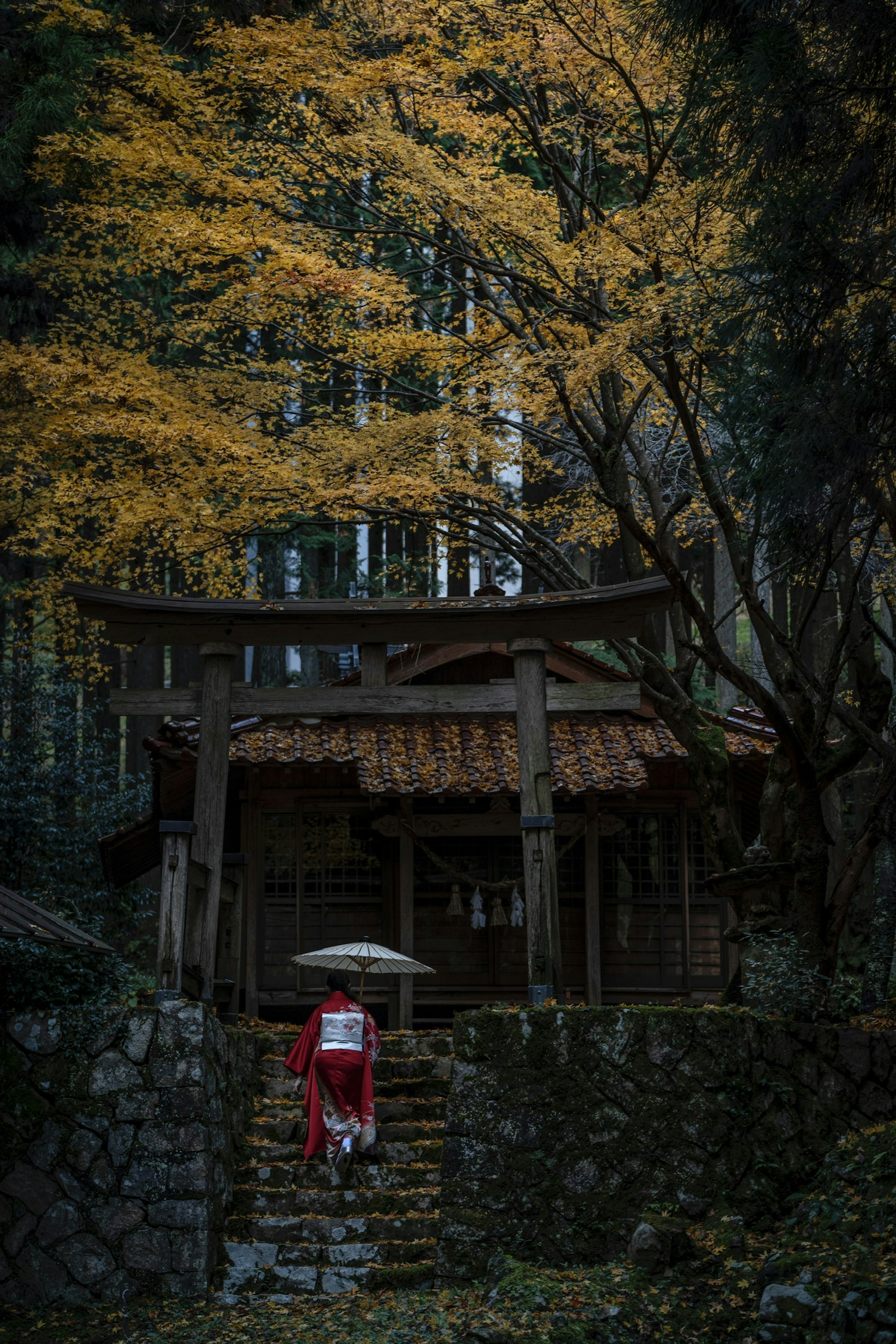 A woman in a red kimono stands under vibrant yellow trees near a traditional Japanese building