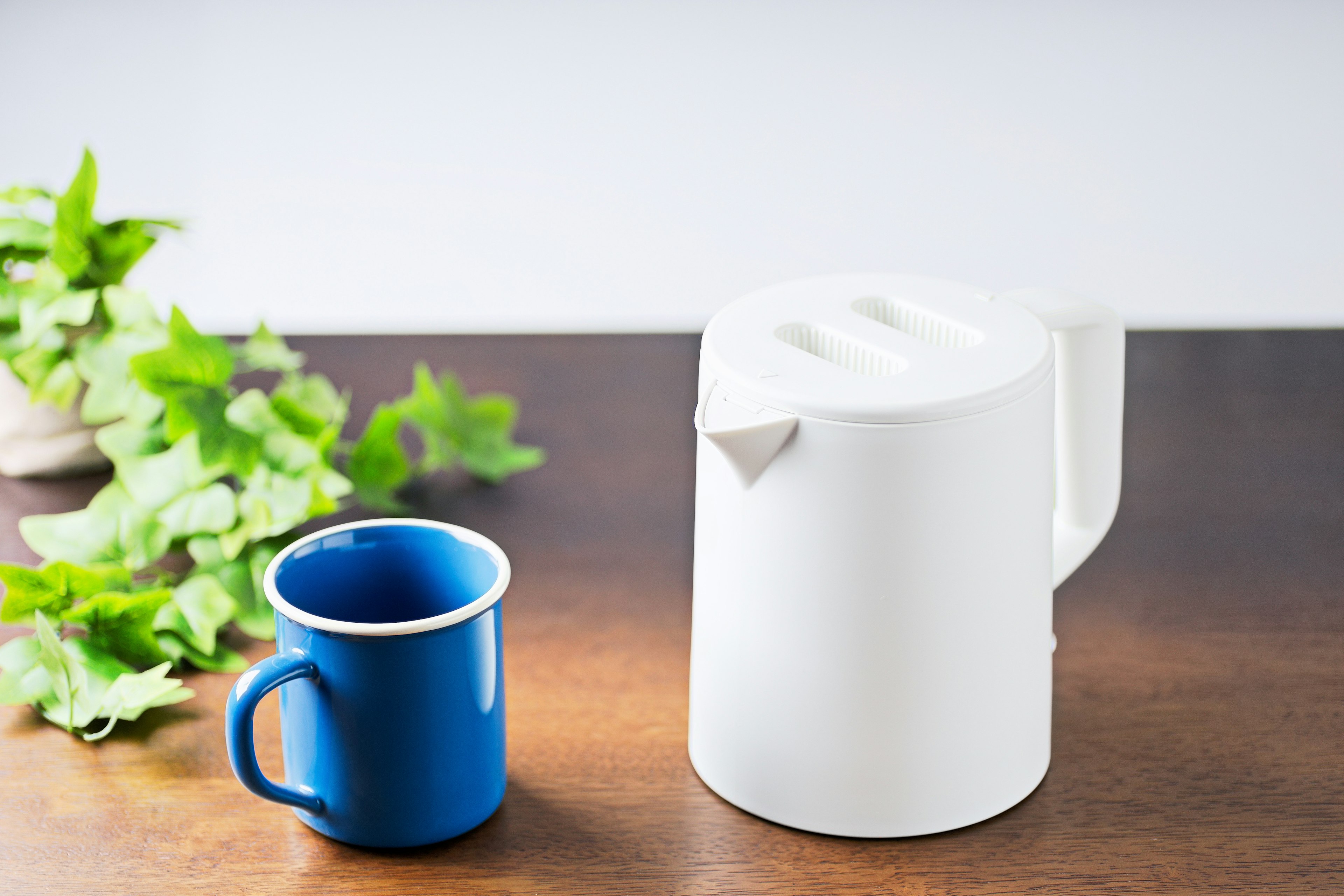 A white teapot and a blue mug are placed on a wooden table with greenery in the background