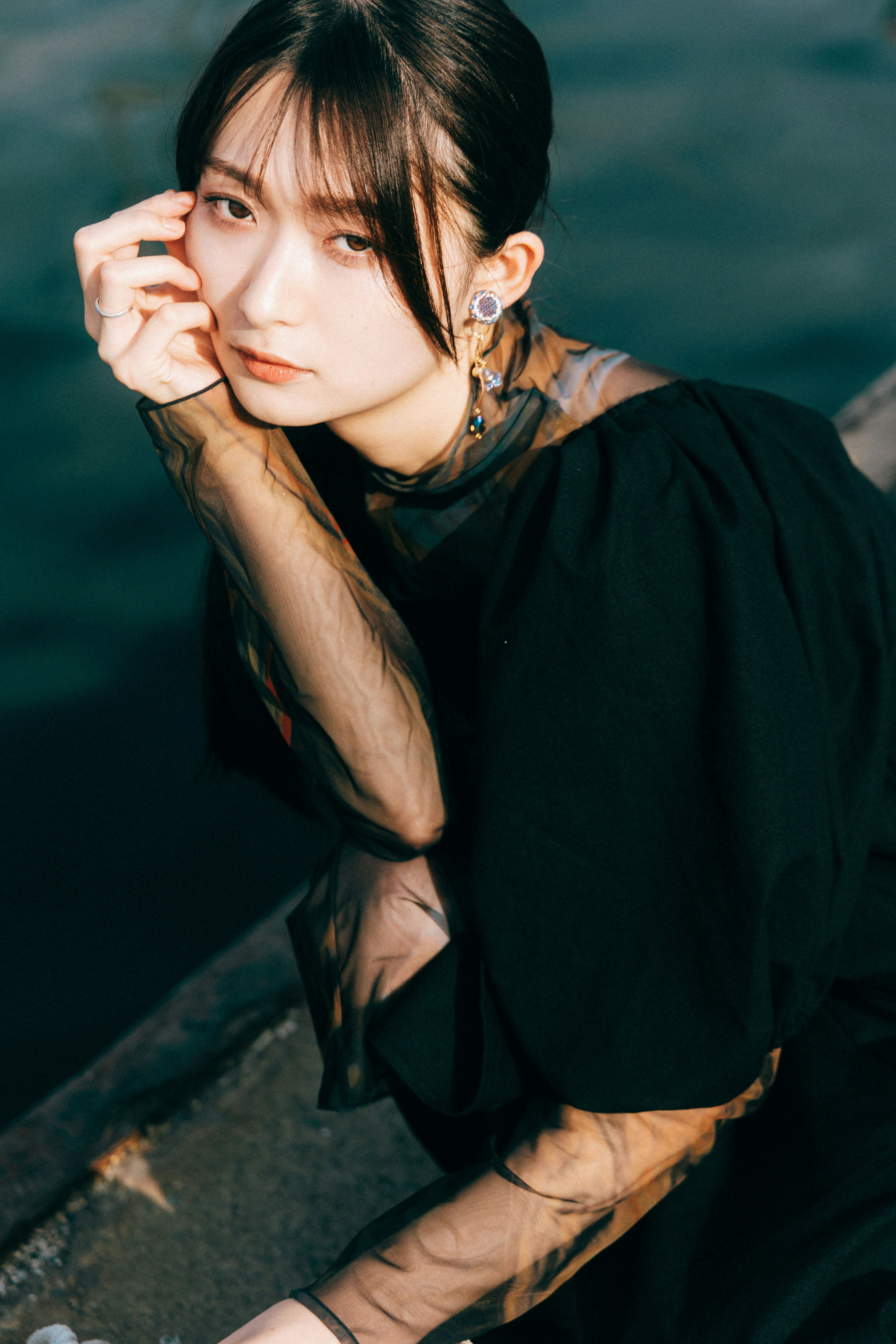 A woman in a black outfit sitting by the water looking contemplative