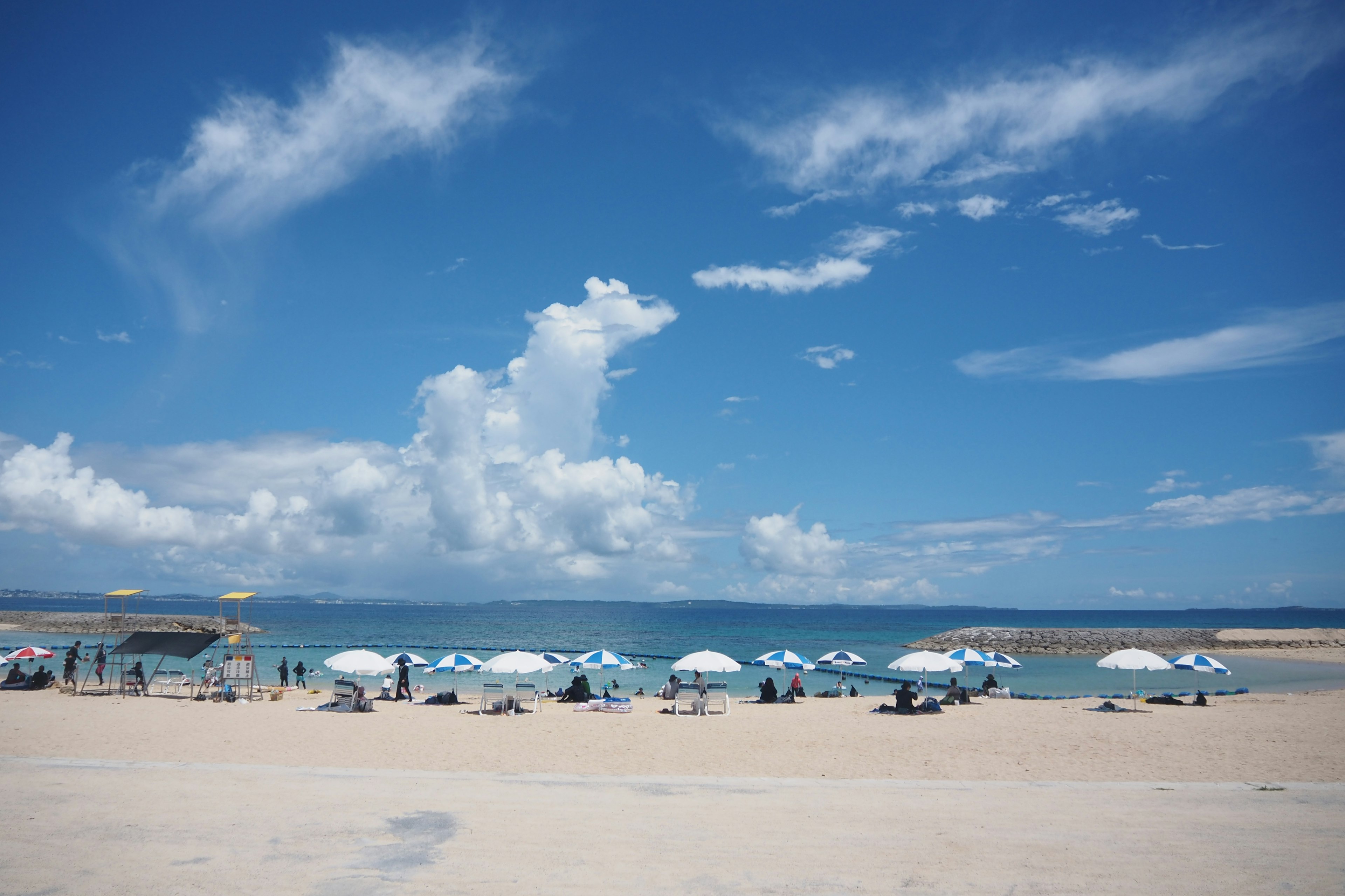 Strandszene mit weißen Sonnenschirmen unter einem blauen Himmel und flauschigen Wolken