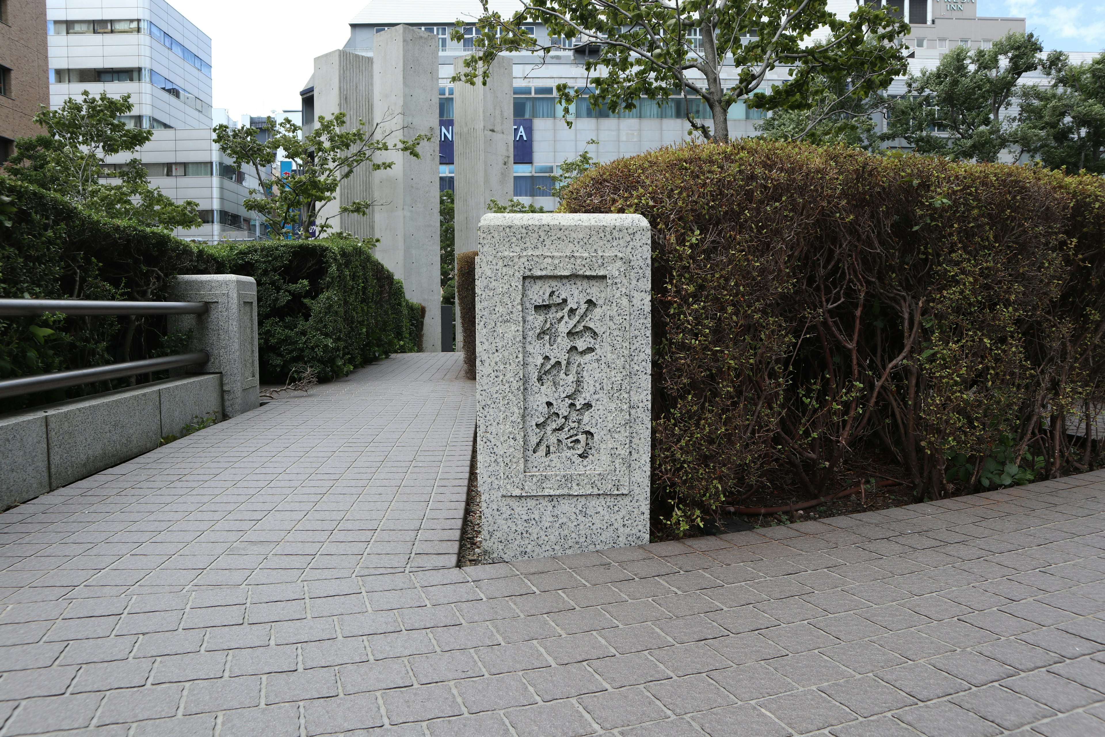 A sidewalk view featuring a stone sign with Japanese characters surrounded by greenery and buildings