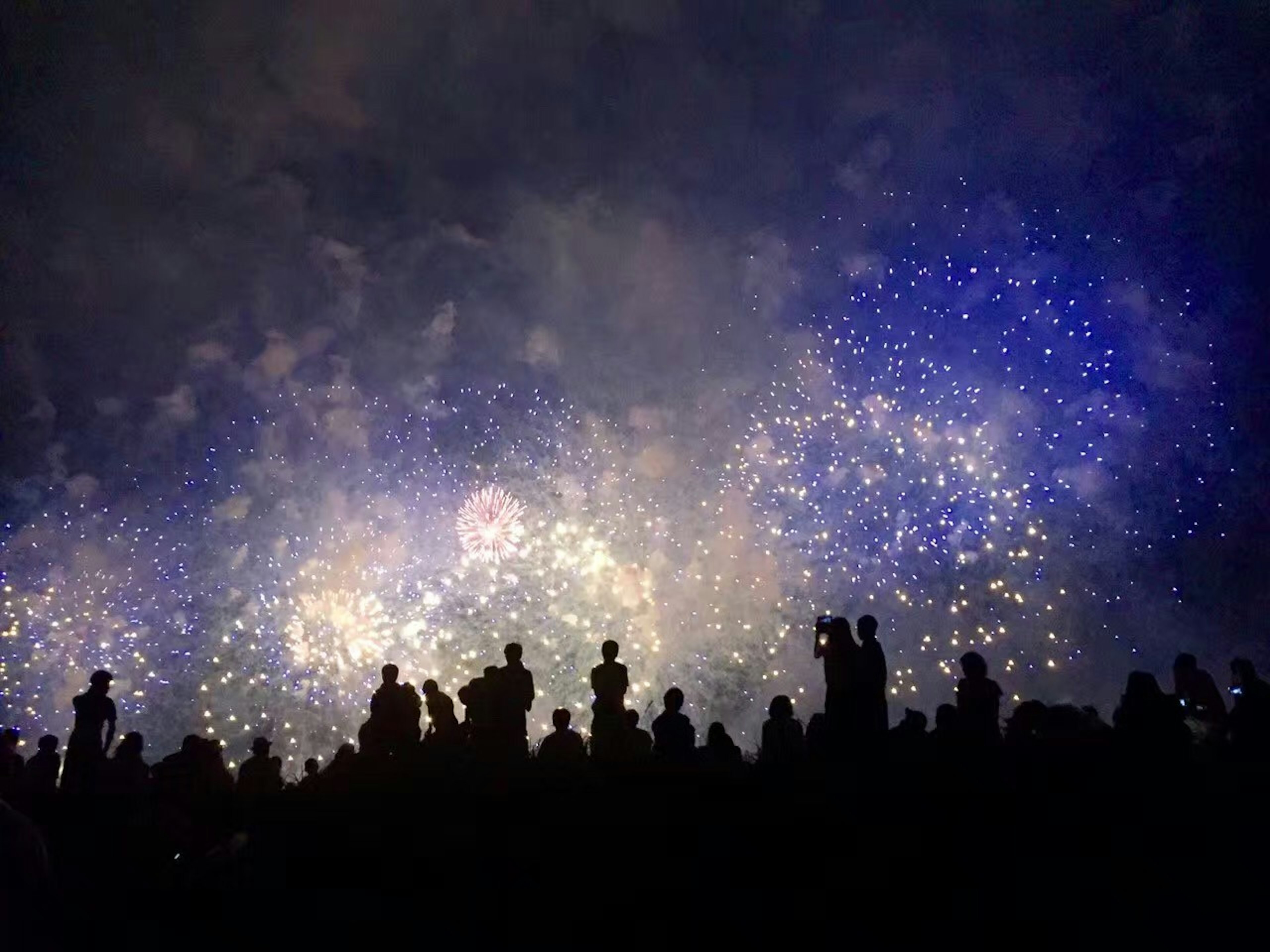 夜空に広がる花火の光と煙の中にシルエットの人々