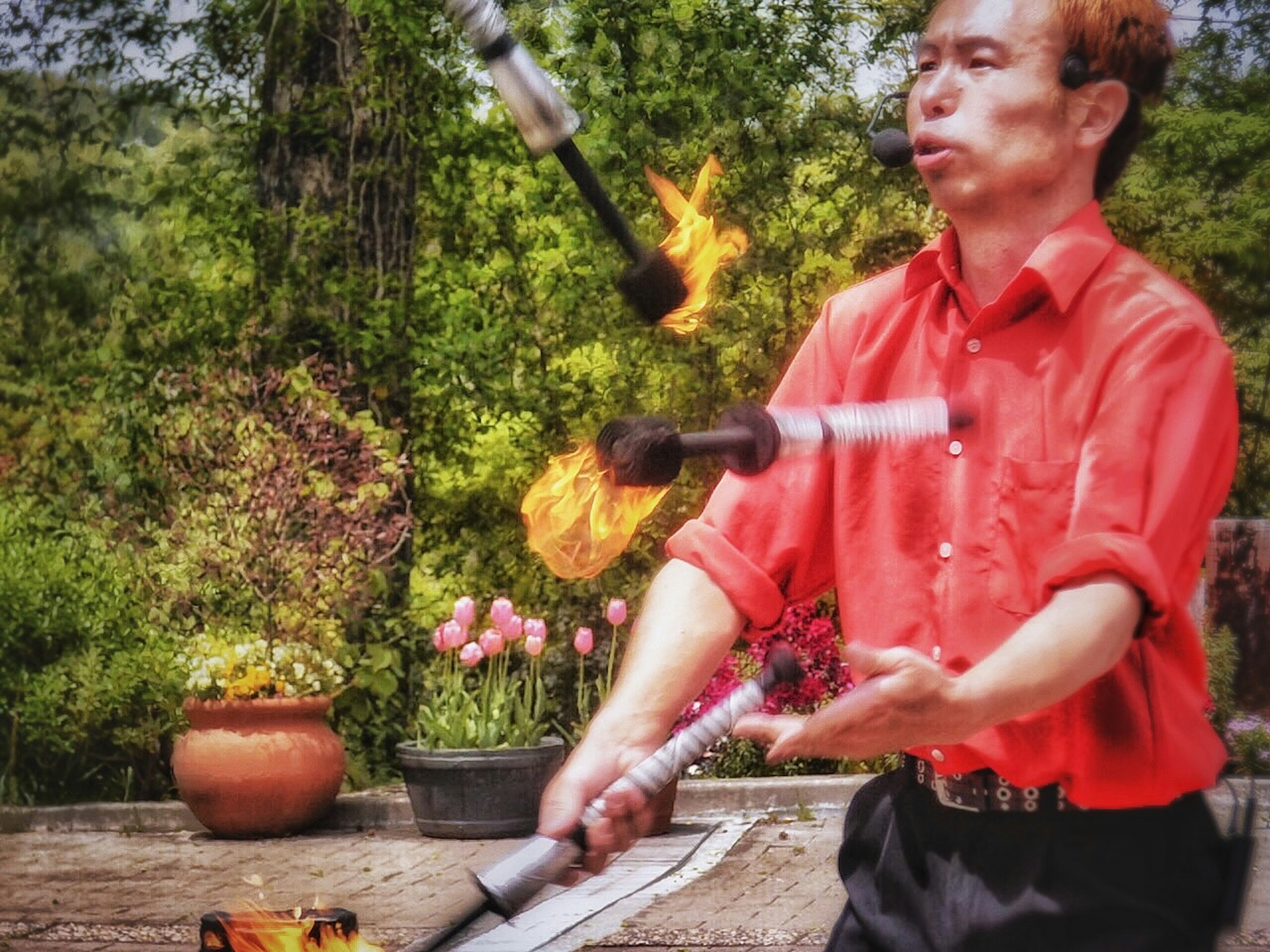 Man juggling with fire wearing a red shirt green trees in the background