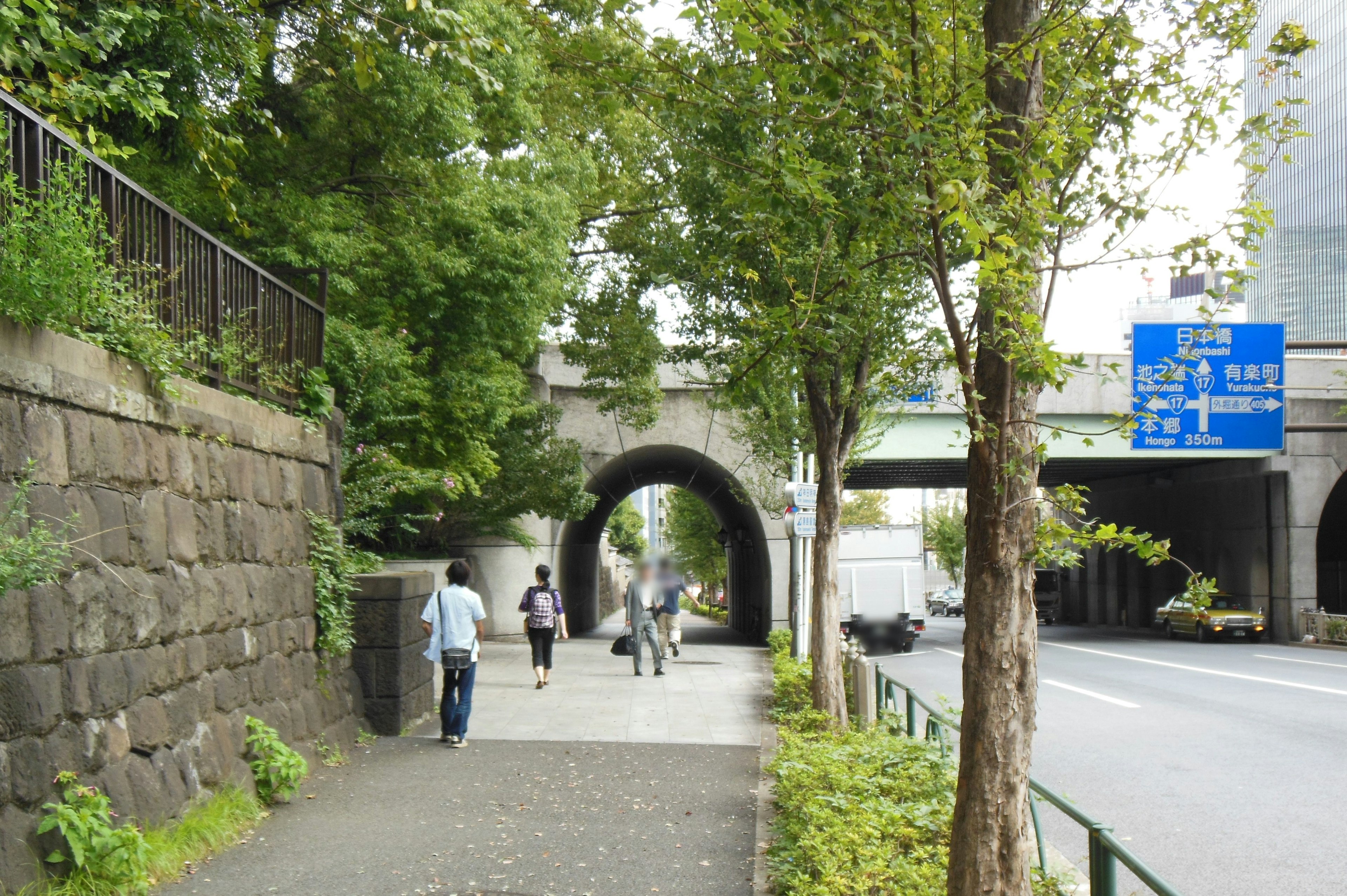 緑の木々に囲まれた歩道とアーチ型の橋の下を歩く人々