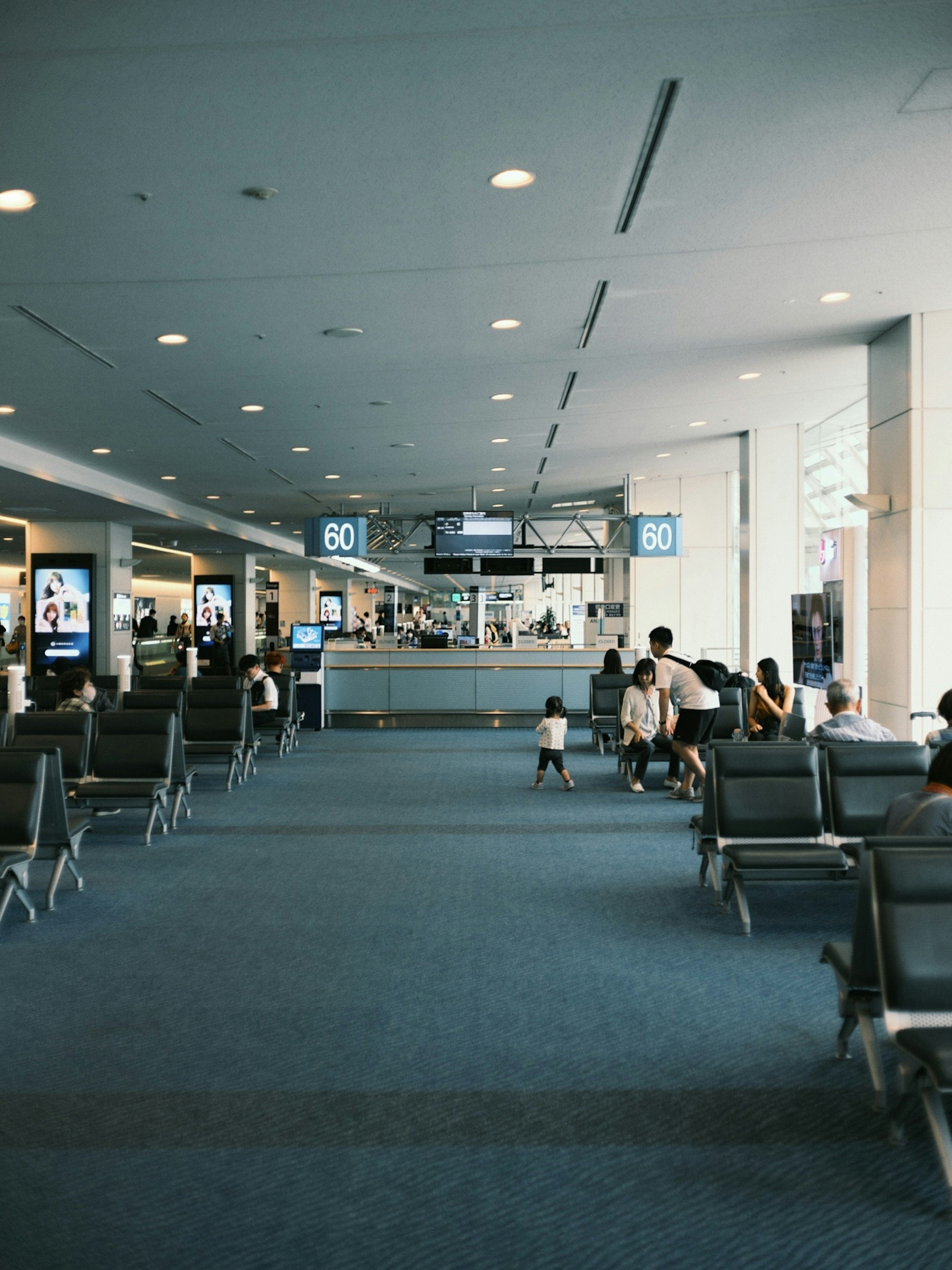 Interior of an airport waiting area Bright lighting and blue carpet Many chairs and people visible