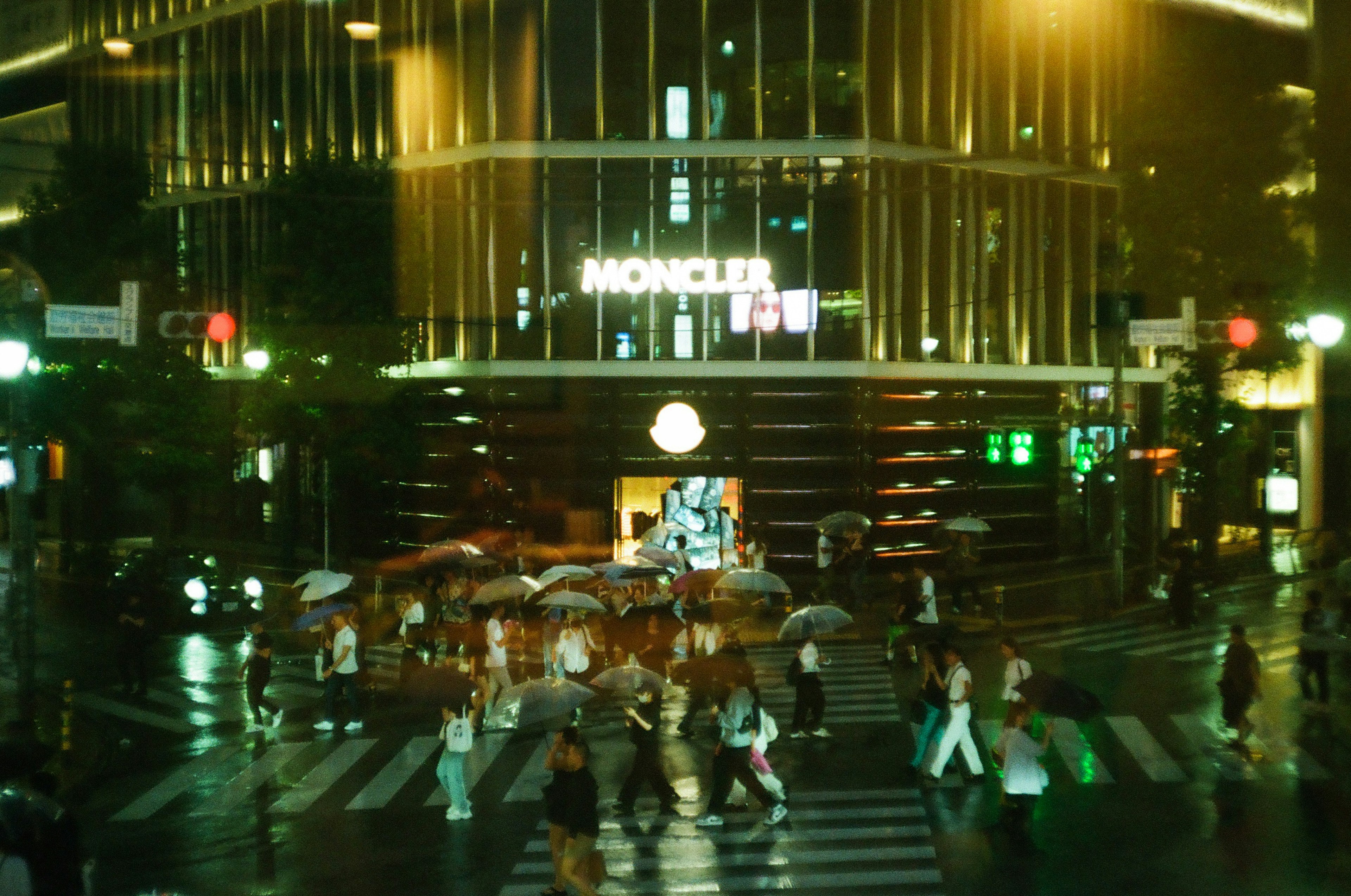 雨の中で傘を持って交差点を渡る人々 モンクレールの看板が目立つ夜の街の景色