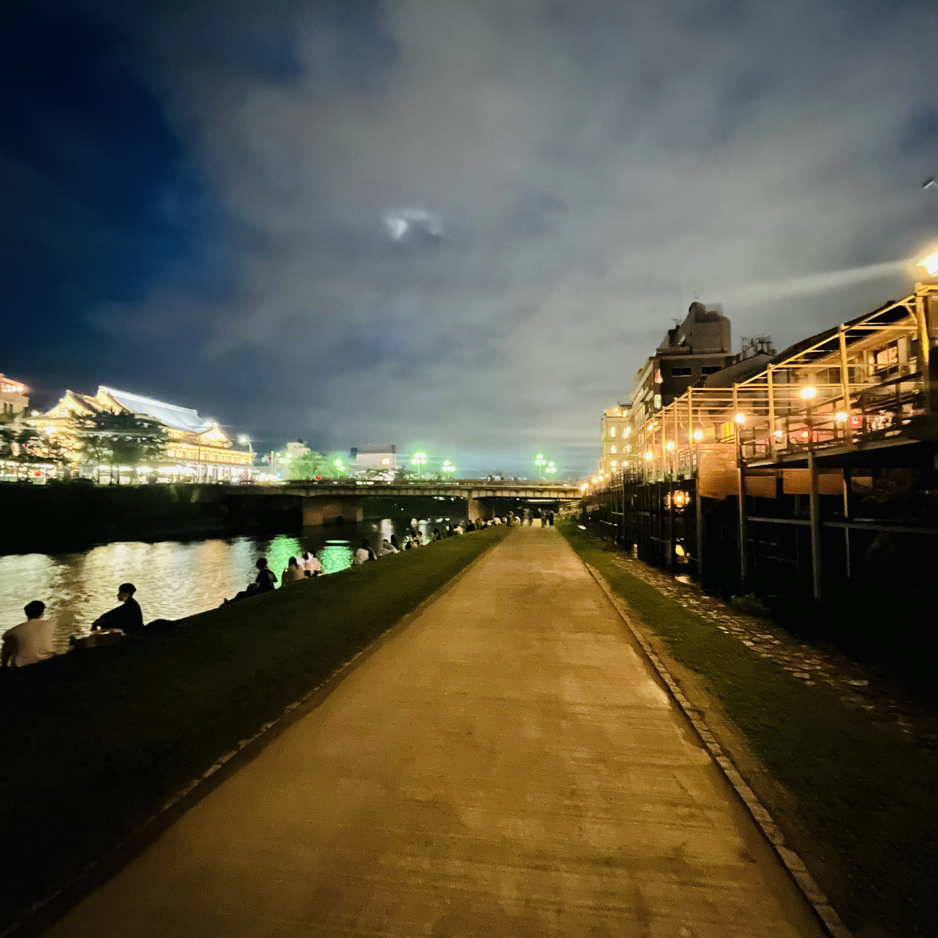 夜の川沿いの散歩道と灯りがともった建物が見える