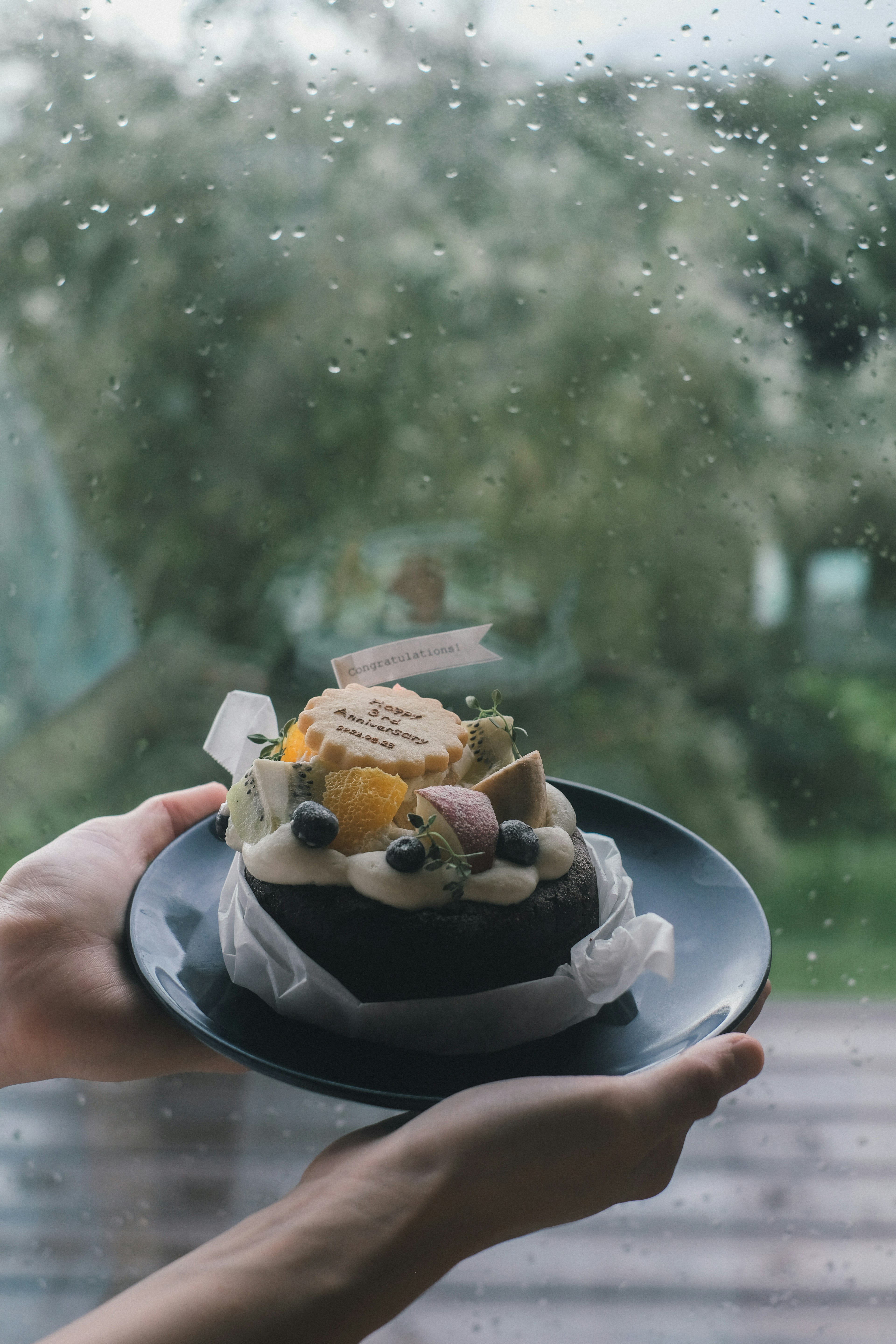 A fruit cake on a black plate held in hands with rain in the background