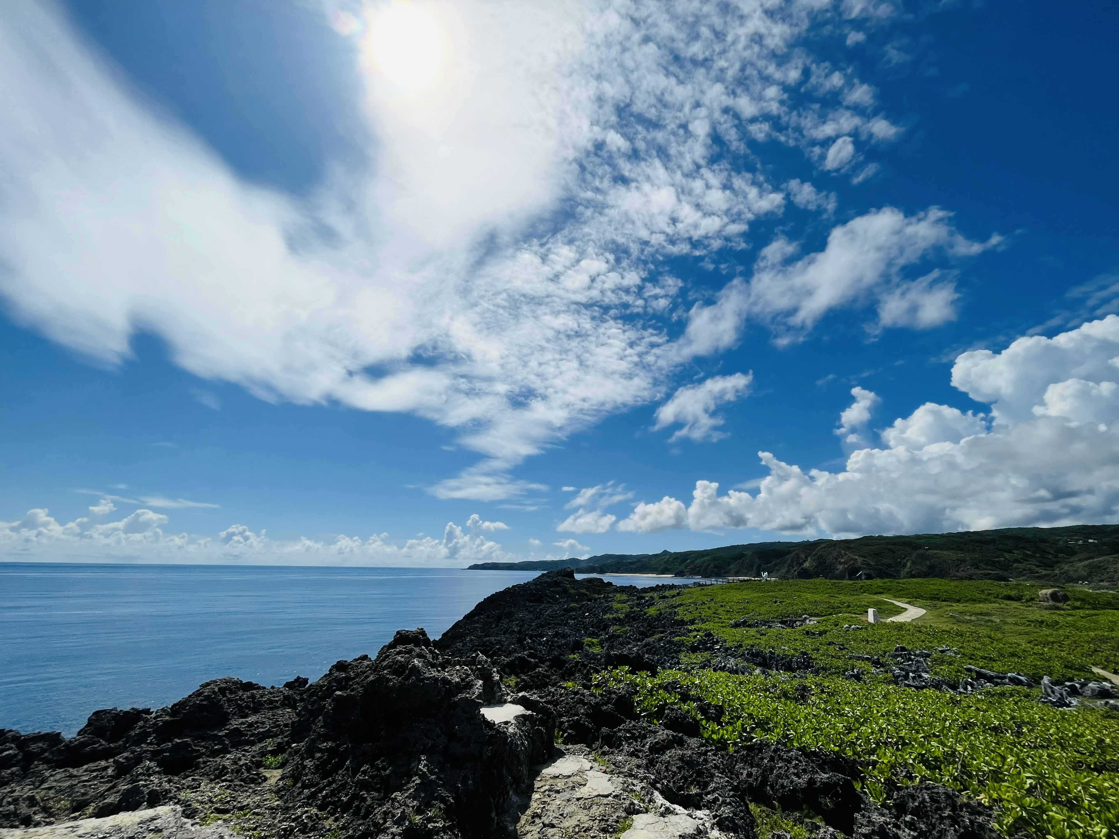 Pemandangan pantai dengan langit biru dan awan putih menampilkan batu dan vegetasi hijau