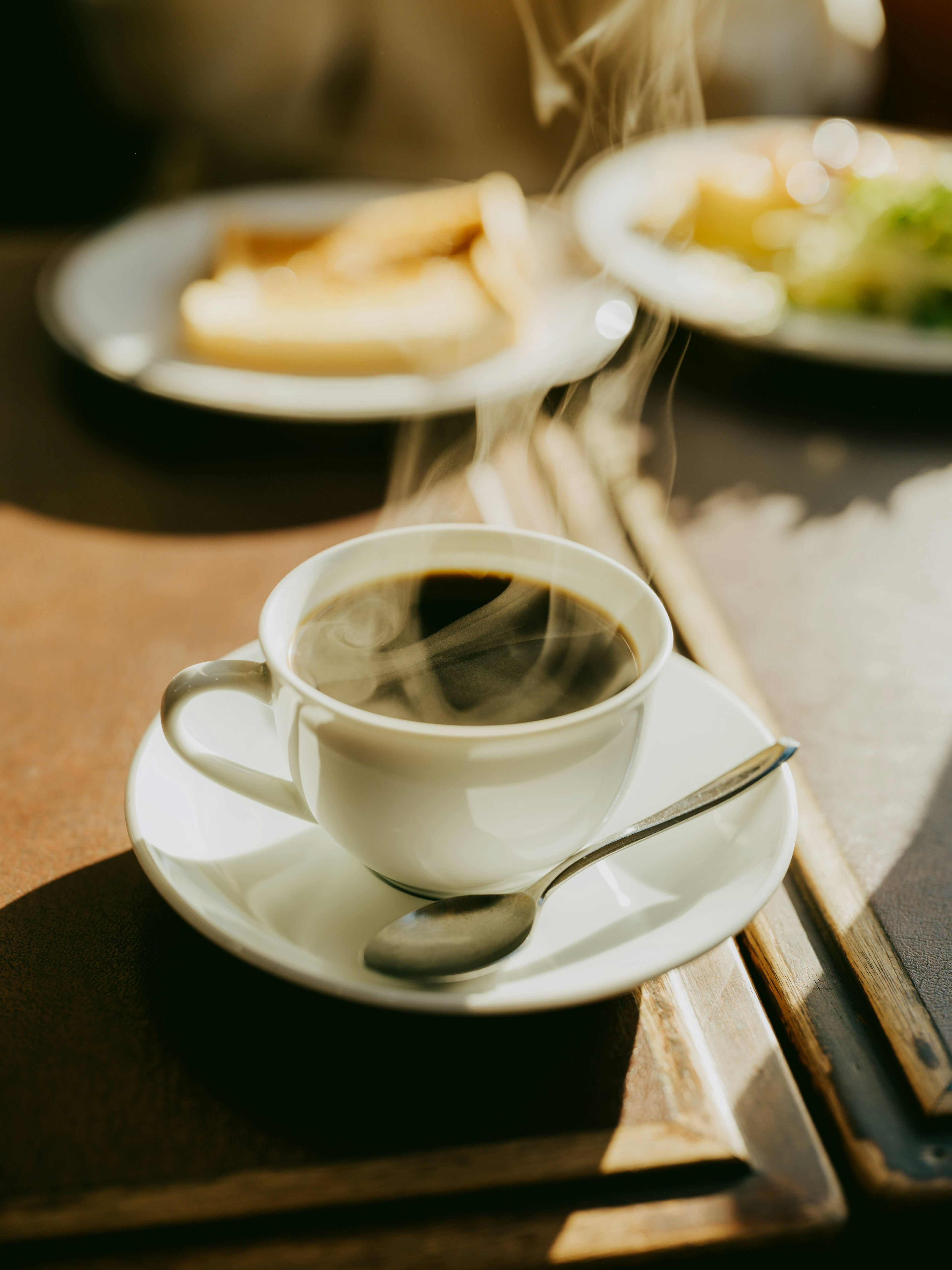 Secangkir kopi panas dalam cangkir putih dengan sendok di samping piring makanan