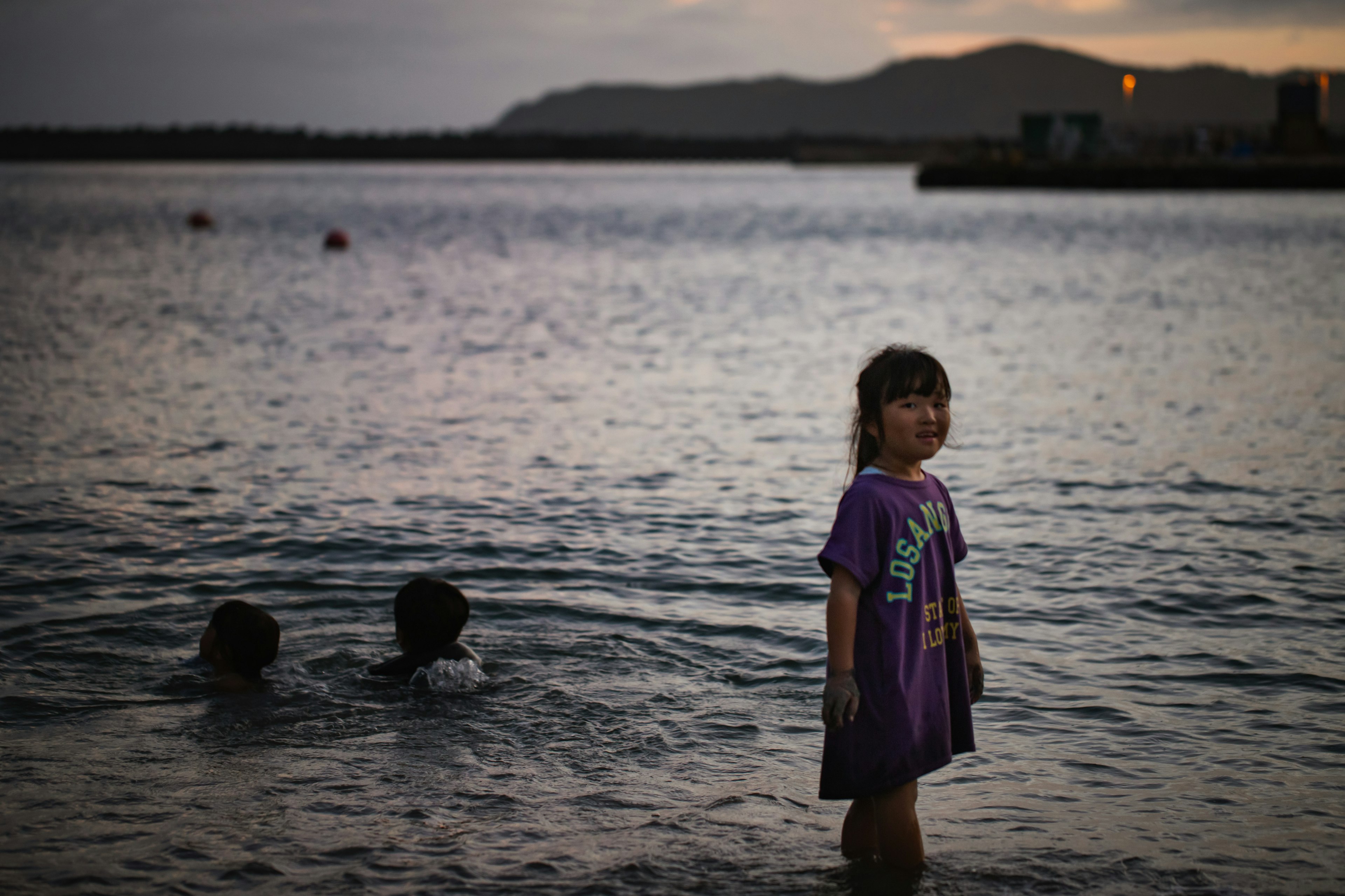 Child standing in water at dusk with two children swimming