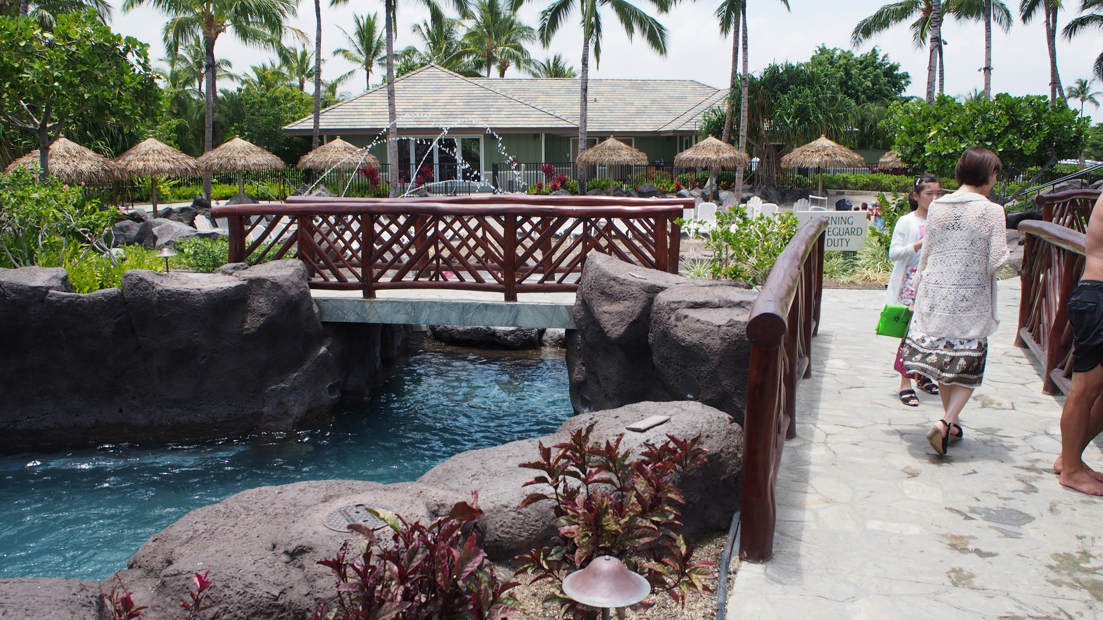 Scenic view of a resort with a wooden bridge and pool surrounded by palm trees and umbrellas