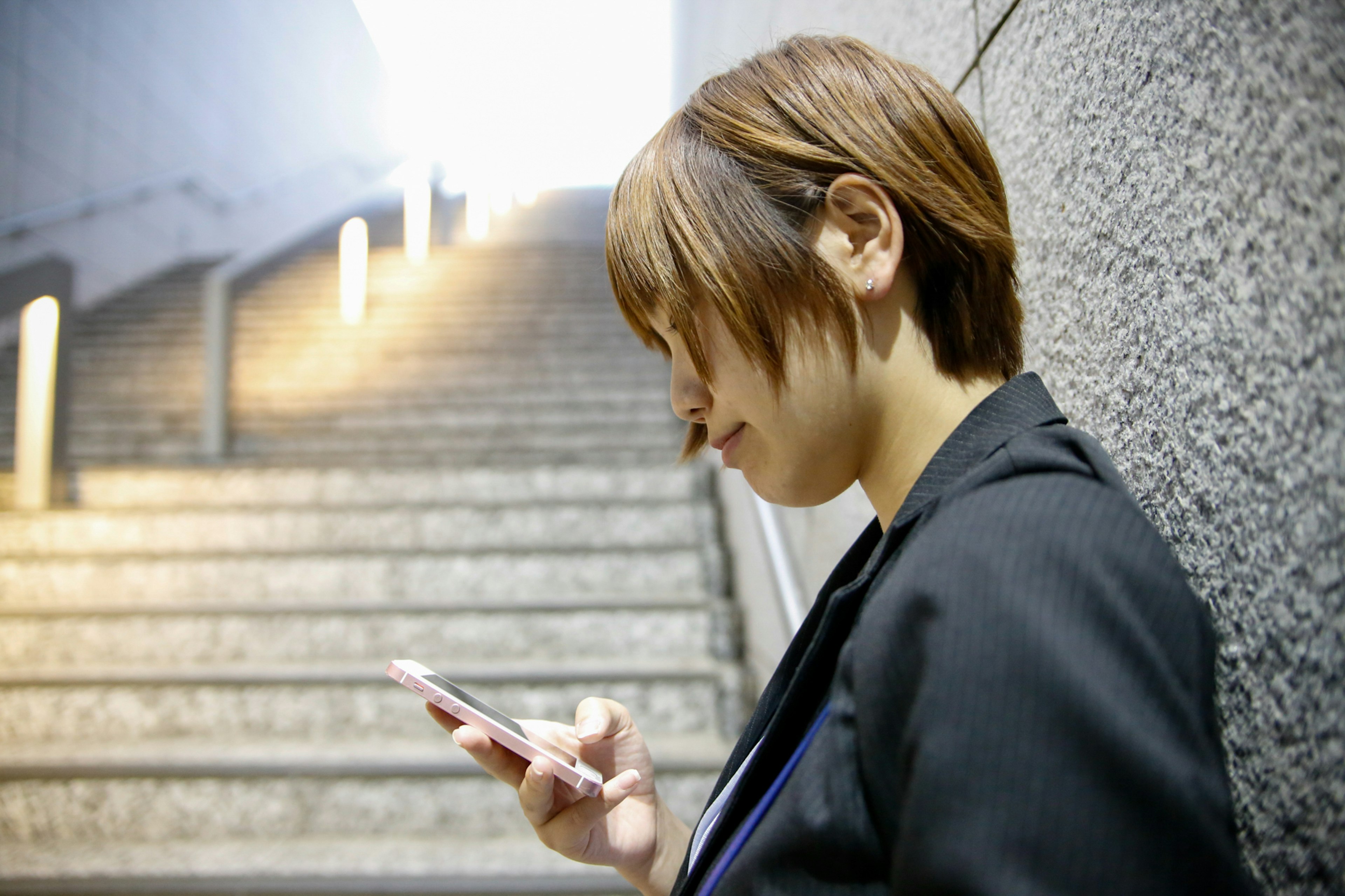 A woman leaning against a staircase wall looking at her smartphone