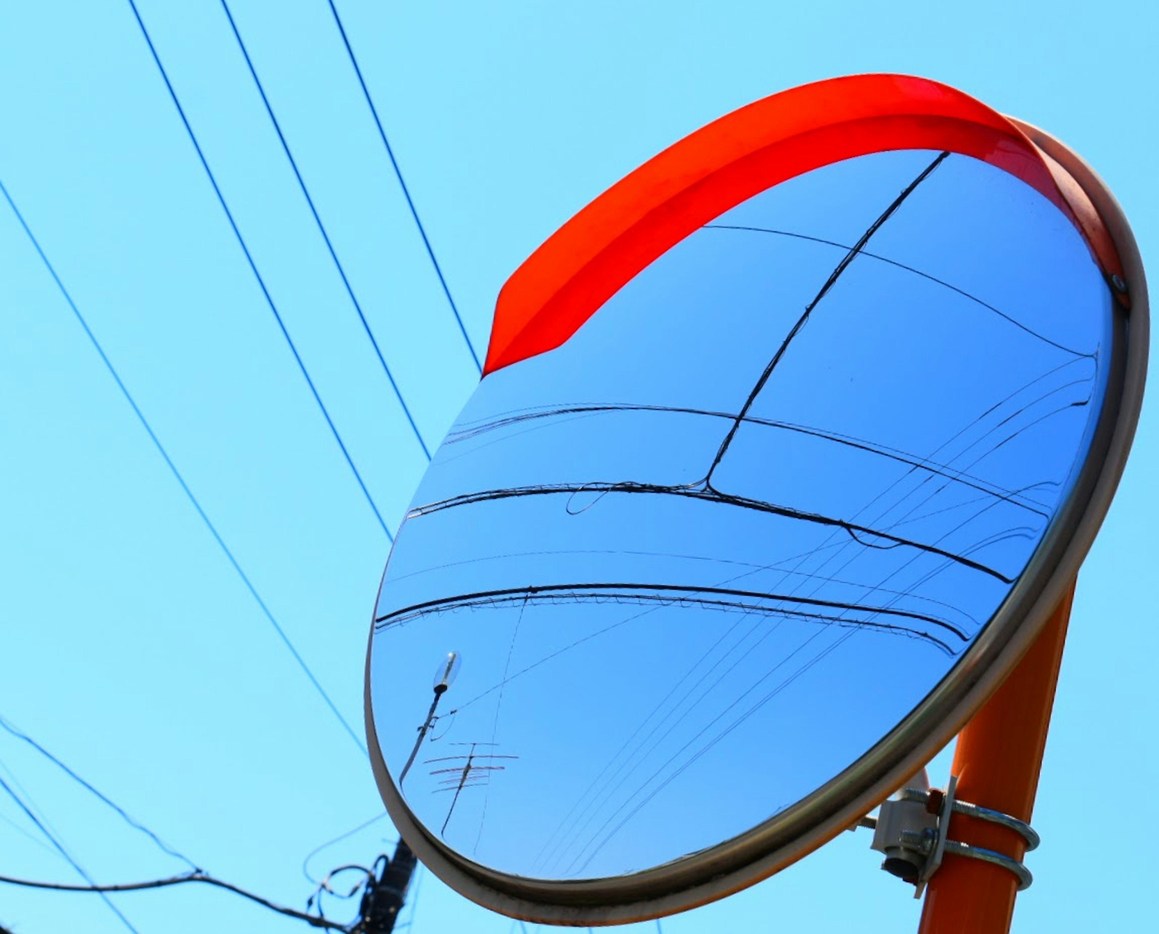 Reflektierender Spiegel mit rotem Rand vor blauem Himmel und Stromleitungen