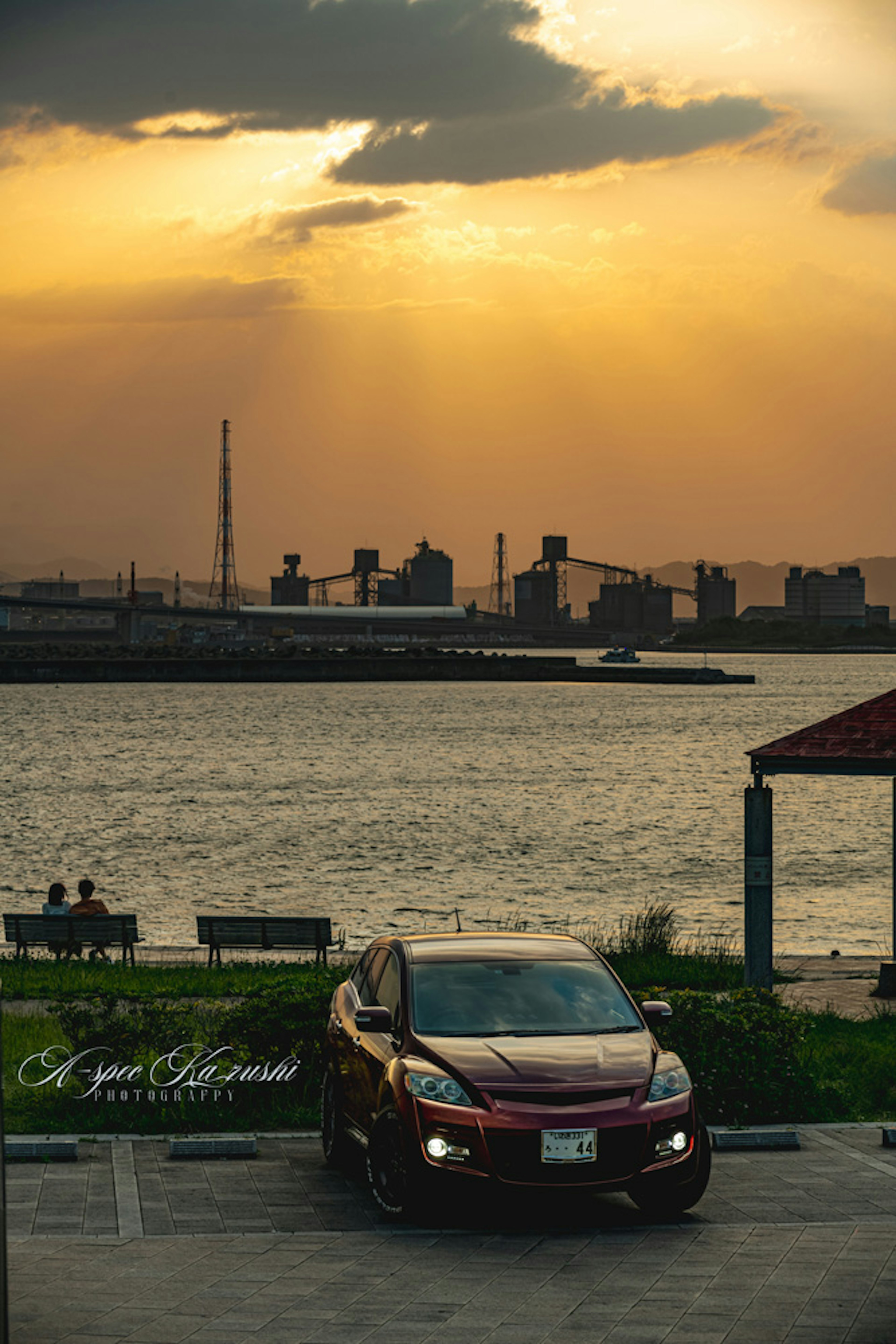 A red car parked by the river at sunset with people in the background