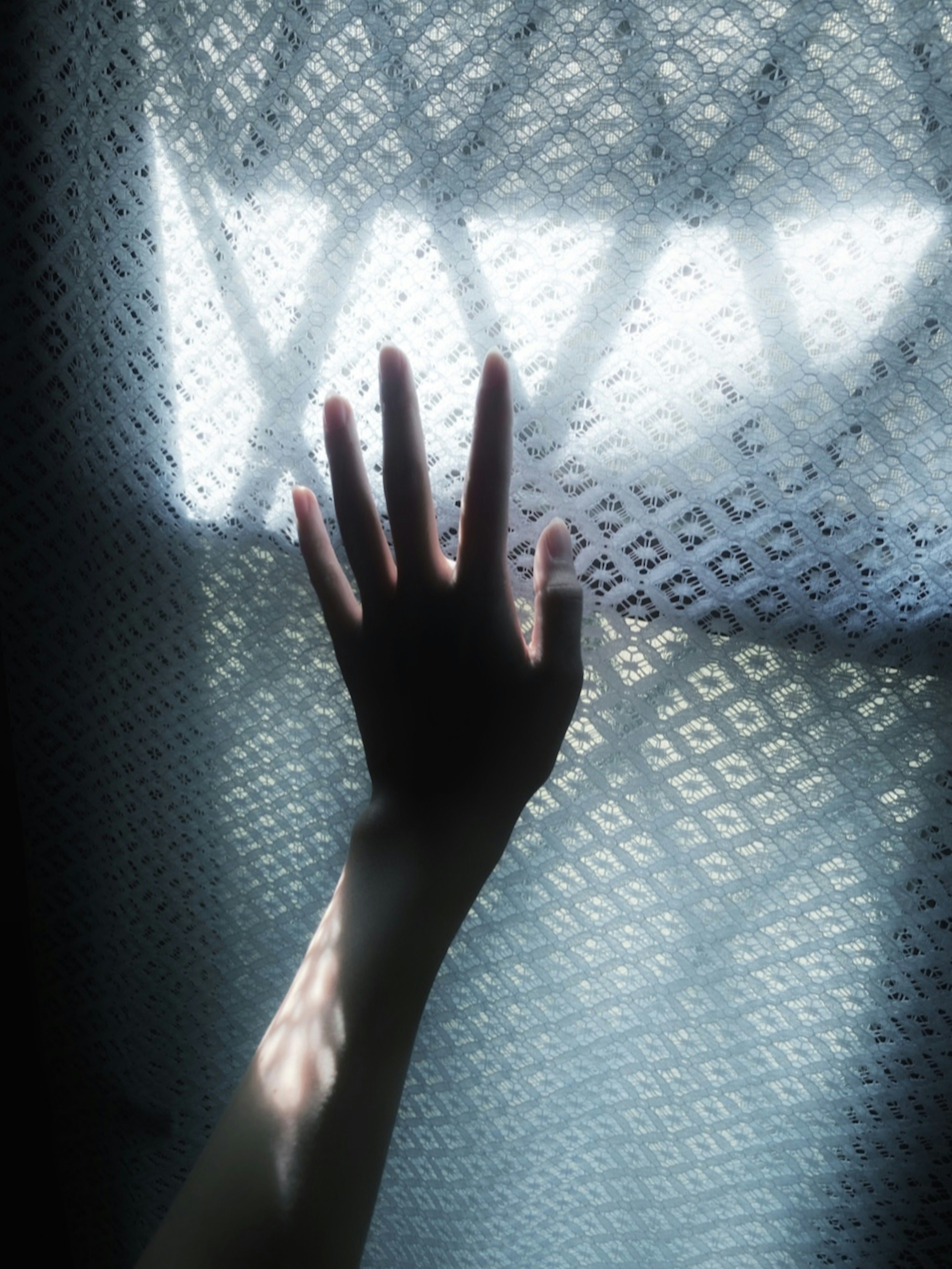 Silhouette of a hand reaching towards light through a patterned curtain