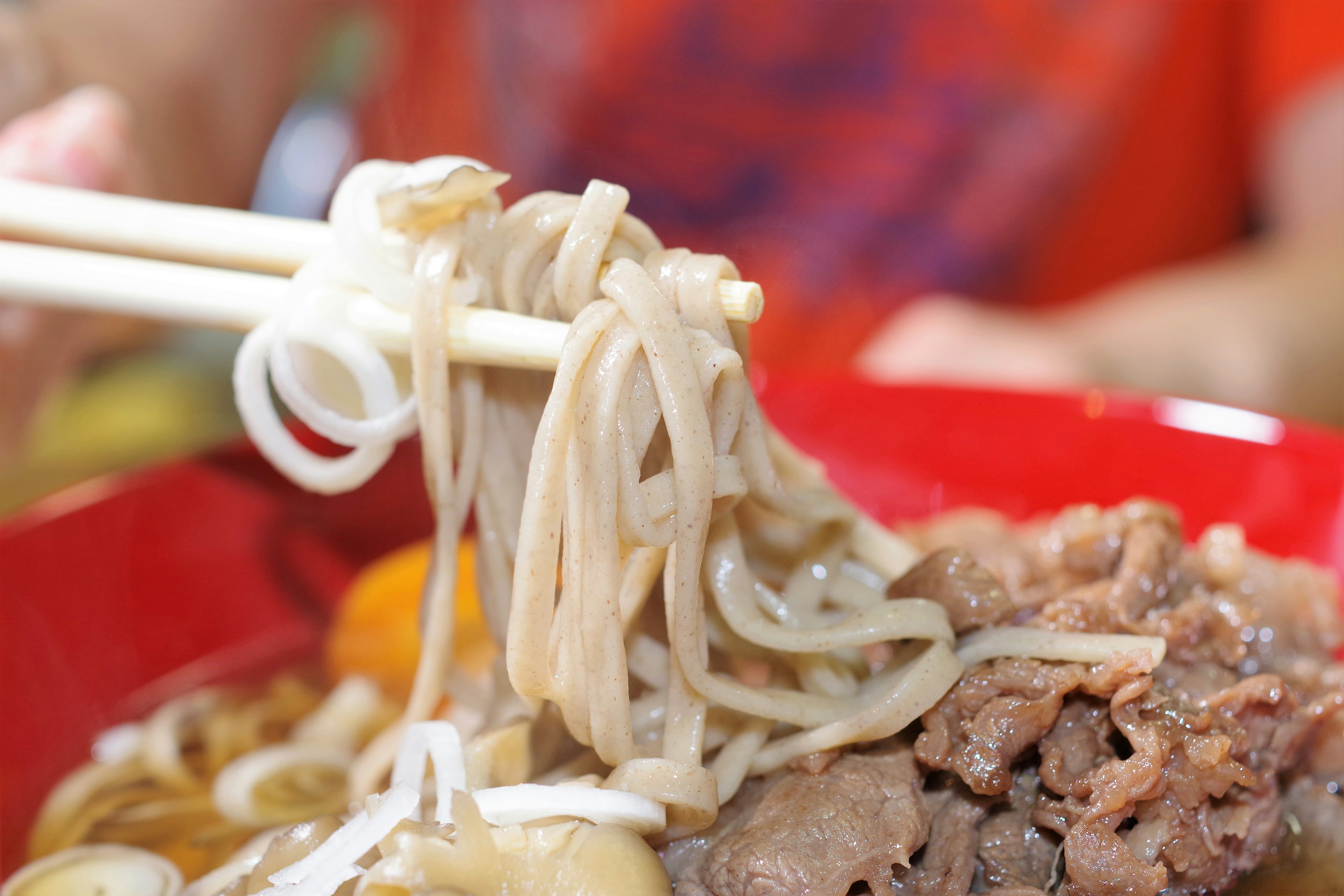 Palillos levantando fideos de un tazón rojo con carne de res y verduras