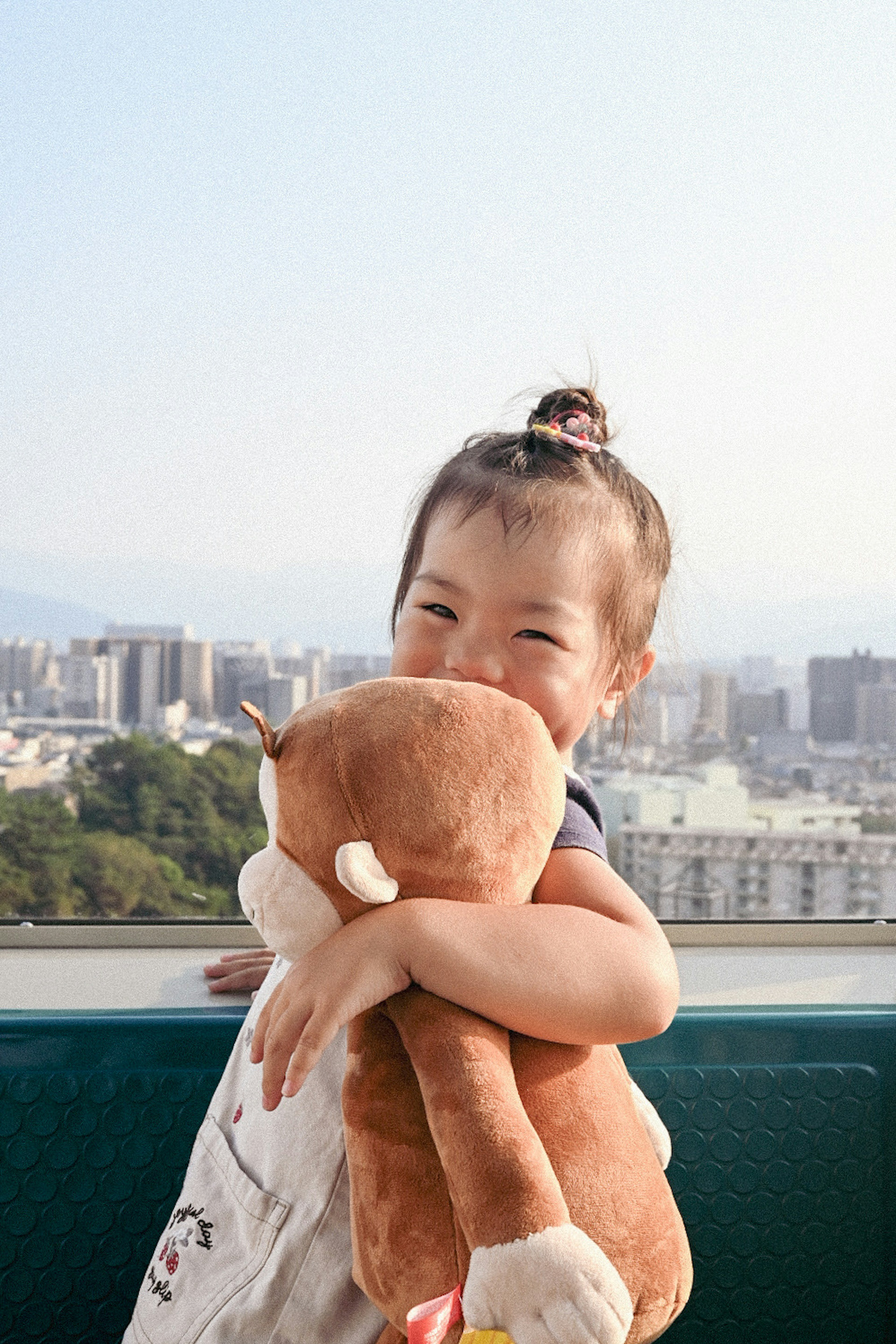 子供が茶色のぬいぐるみを抱きしめている笑顔の写真 背景には都市の風景が広がっている