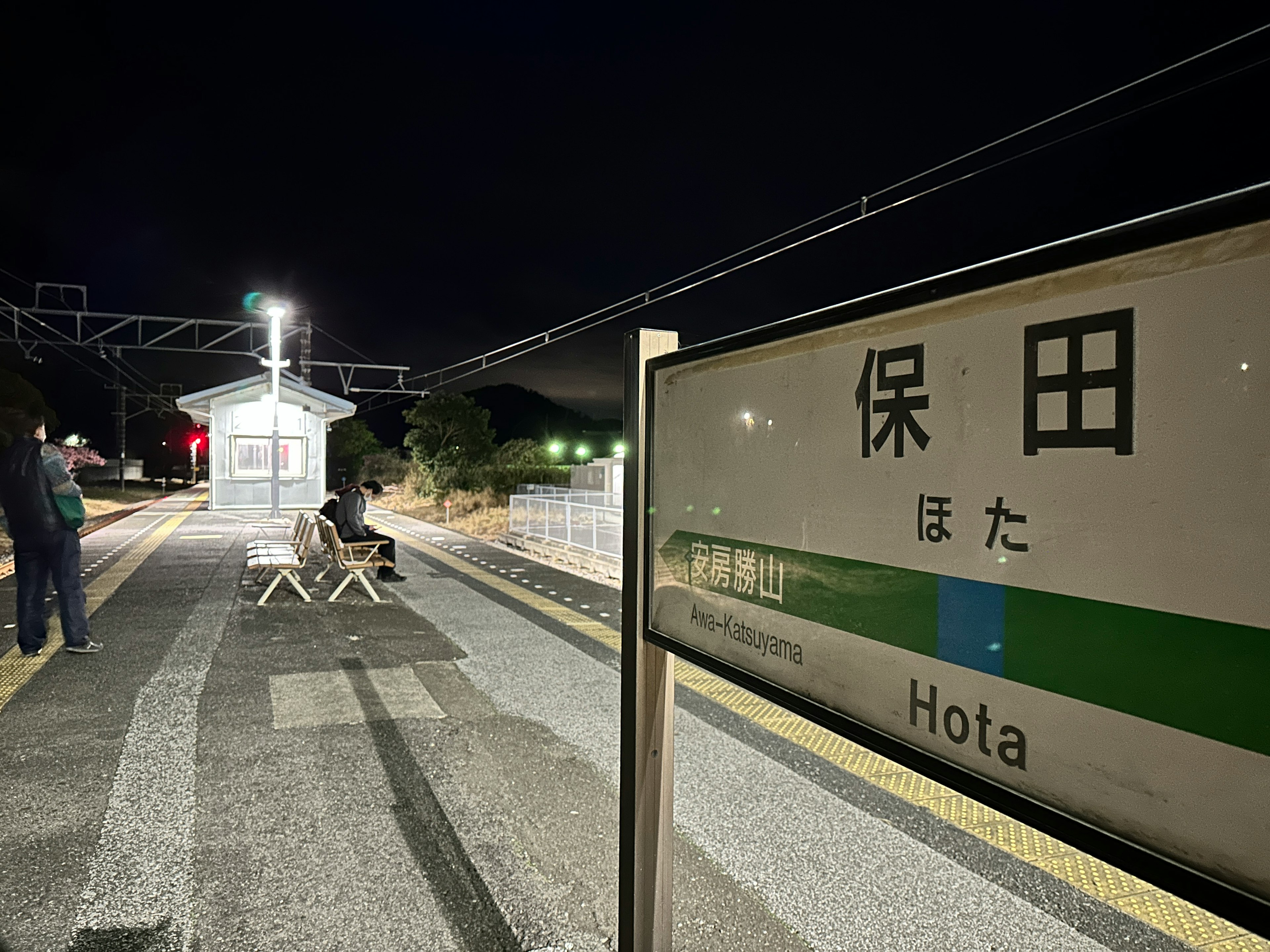 夜の保田駅のプラットフォームに立つ駅名標と人