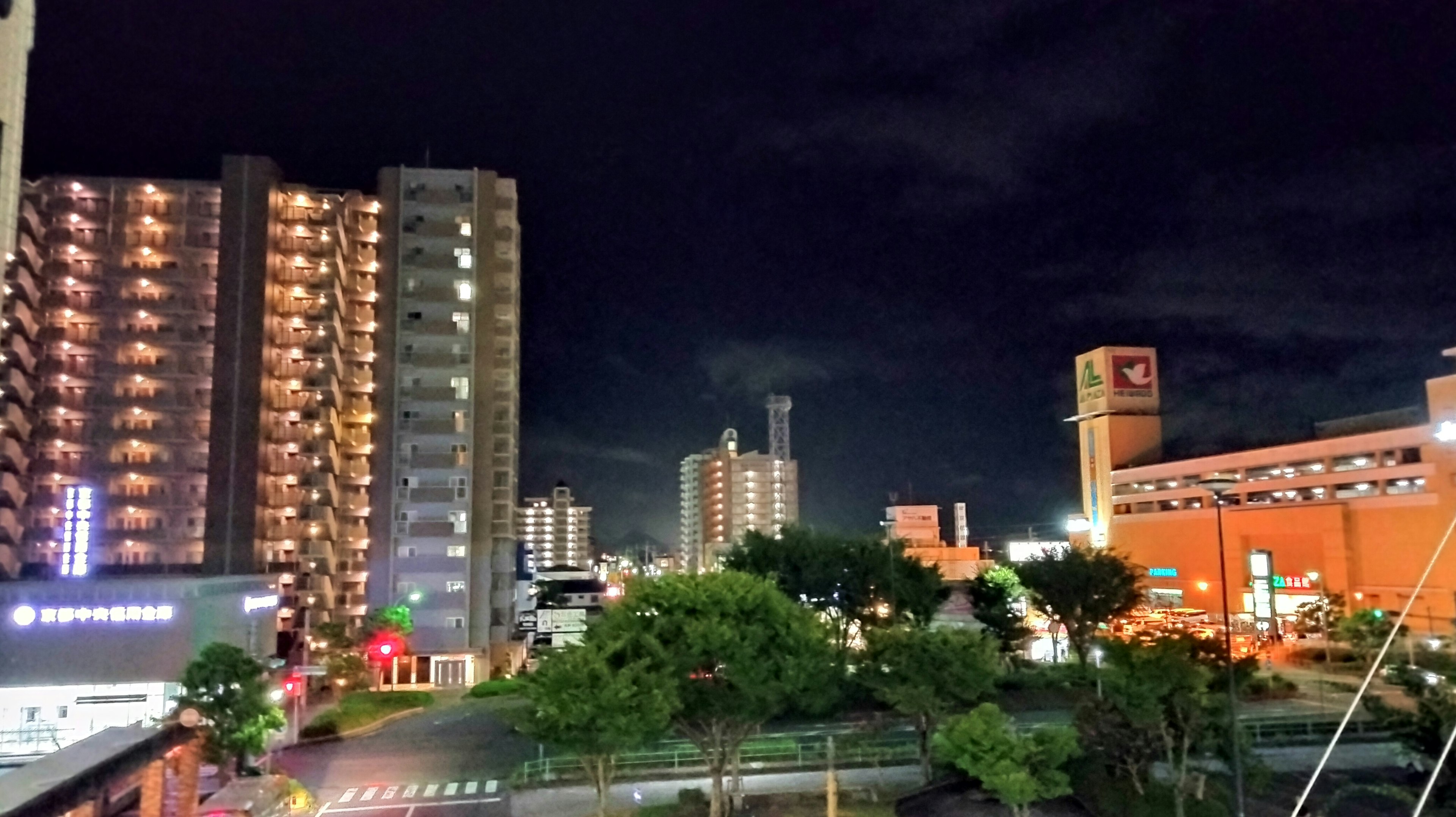 Night cityscape featuring tall buildings and green trees under a dark sky