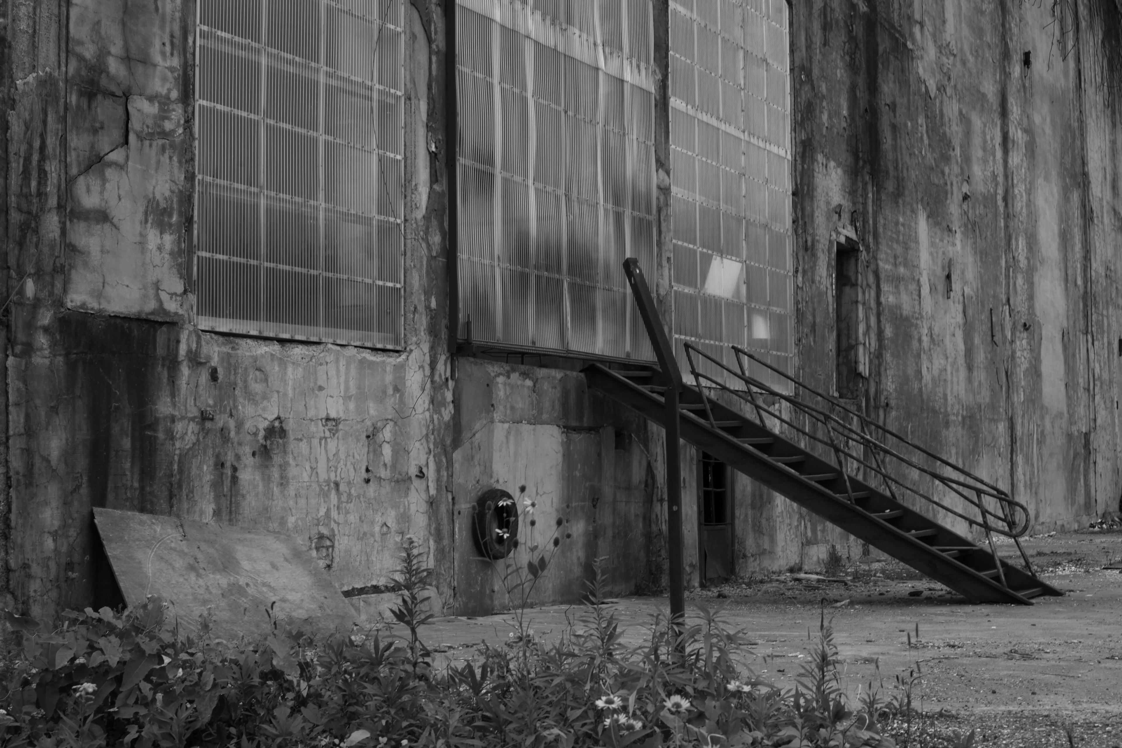 Extérieur d'une vieille usine avec des escaliers rouillés de grandes fenêtres et des murs usés envahis par l'herbe