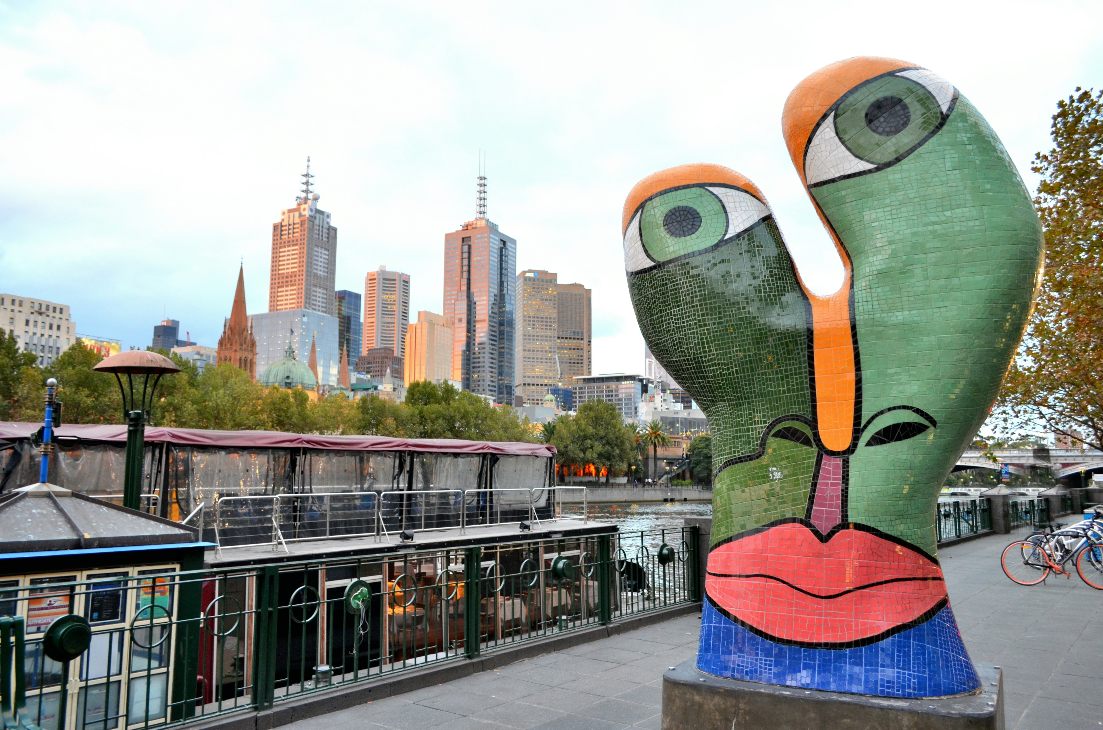 Patung berwarna-warni dengan motif wajah di latar belakang kota Melbourne