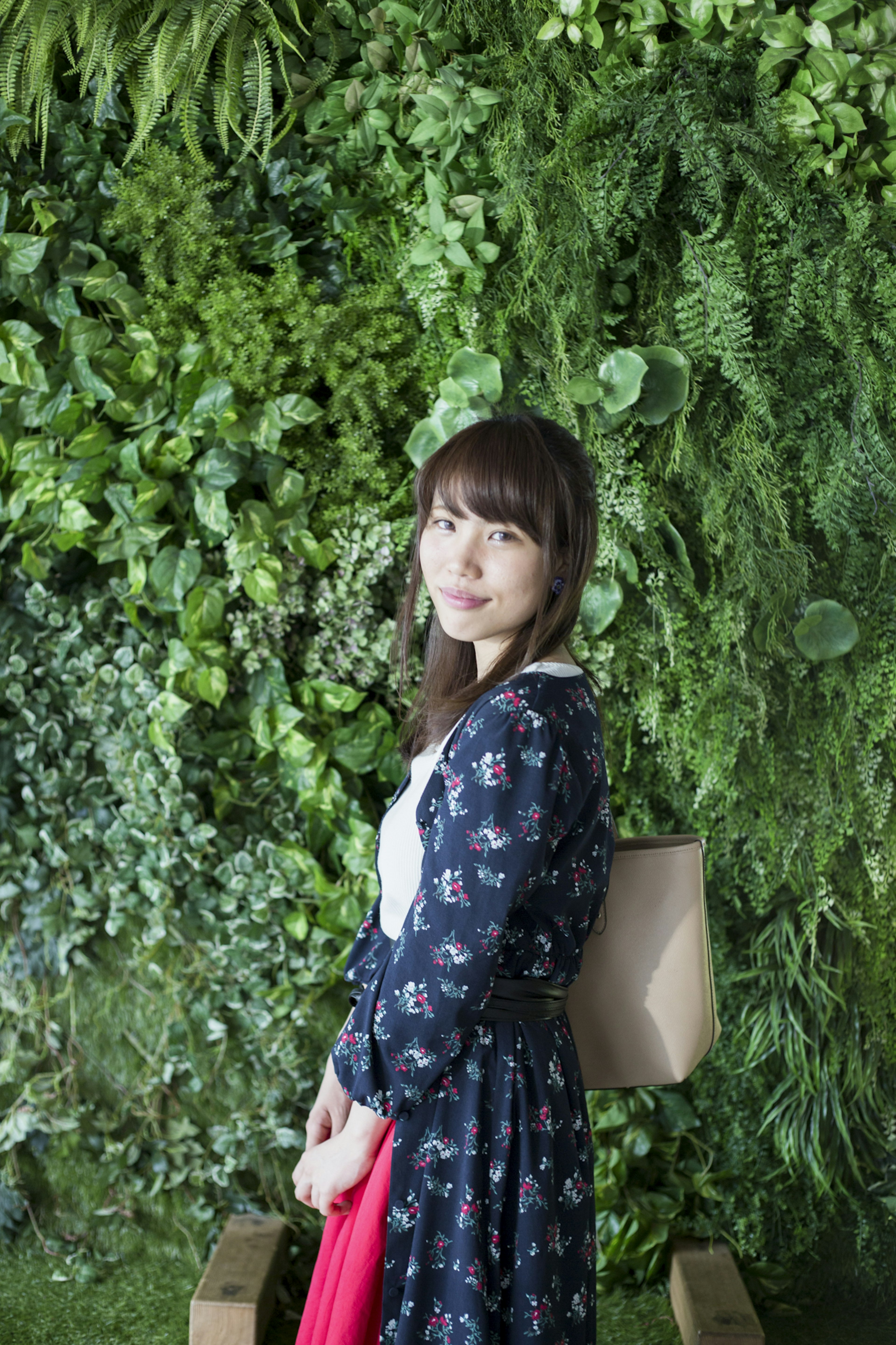 Portrait of a woman standing in front of a green wall wearing a floral dress and holding a bag