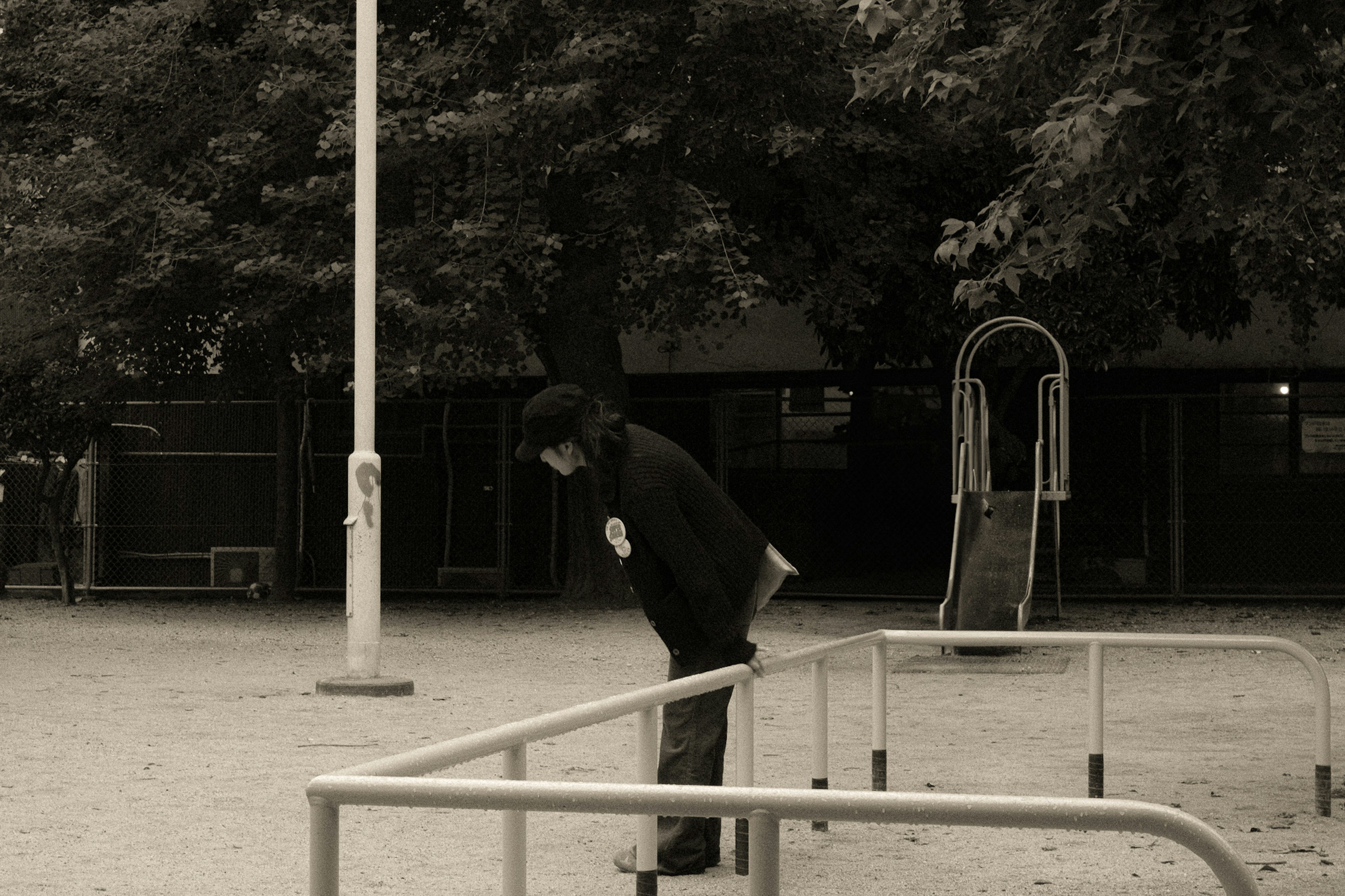 Persona apoyada en una barandilla de parque en tonos de blanco y negro con árboles de fondo