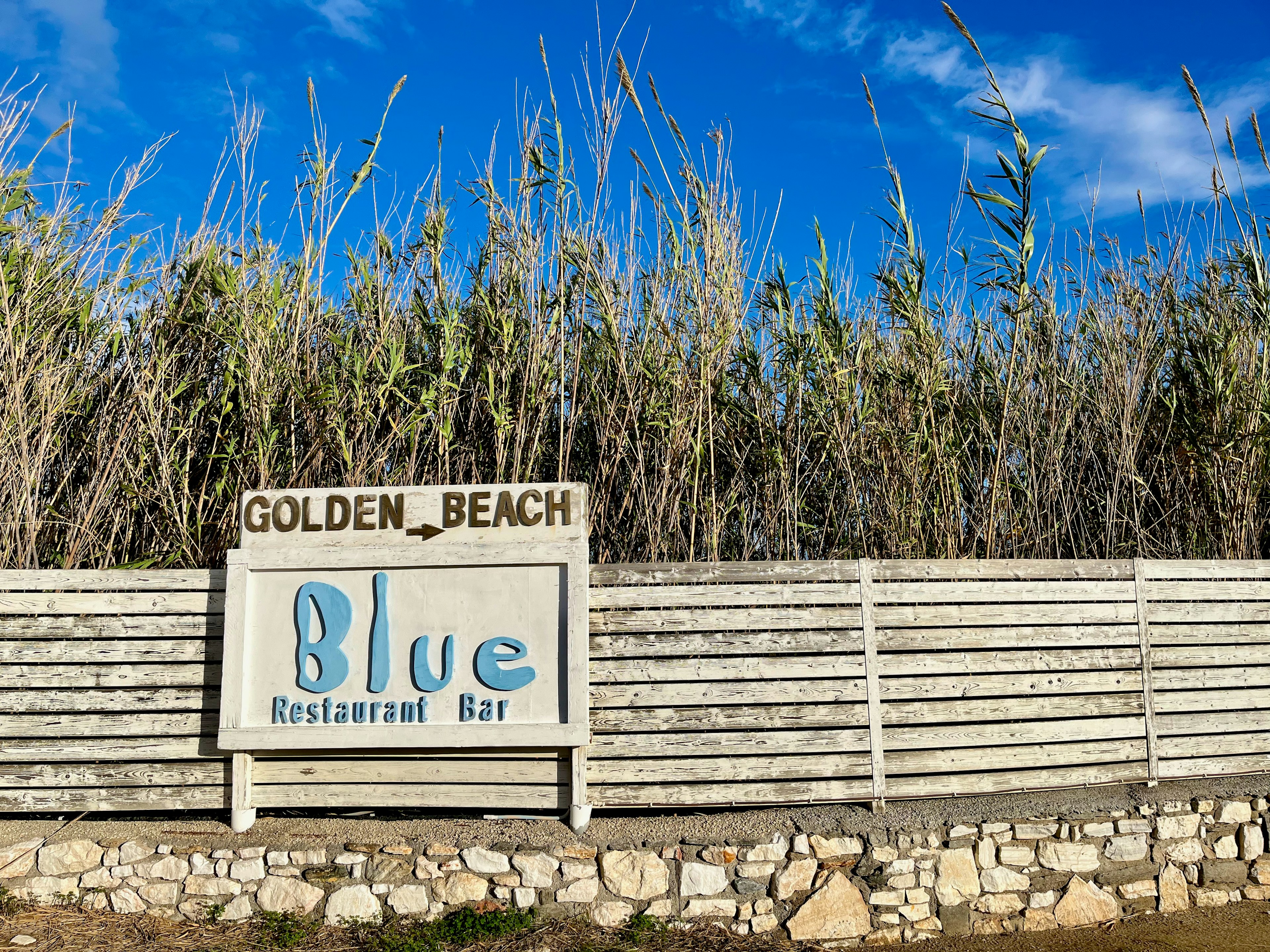 Schild des Blue Restaurant Bar am Golden Beach umgeben von hohem Gras