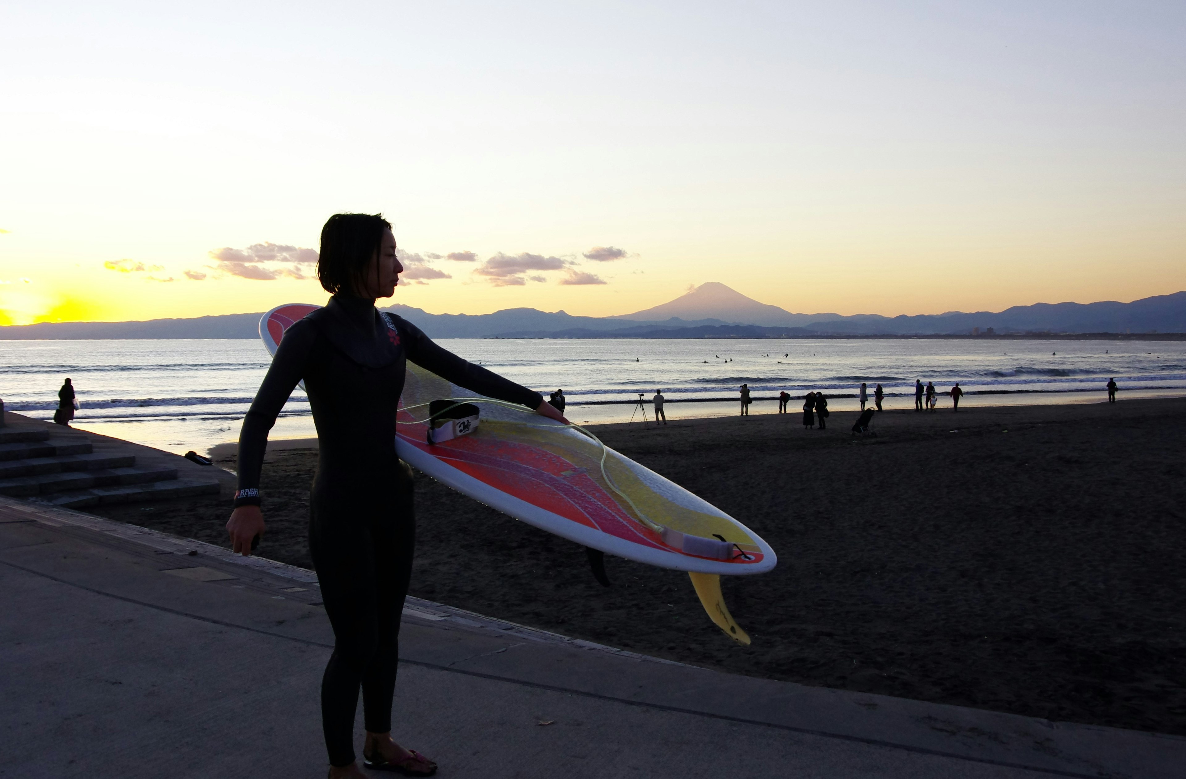 女性在日落背景下手持冲浪板，背后有山