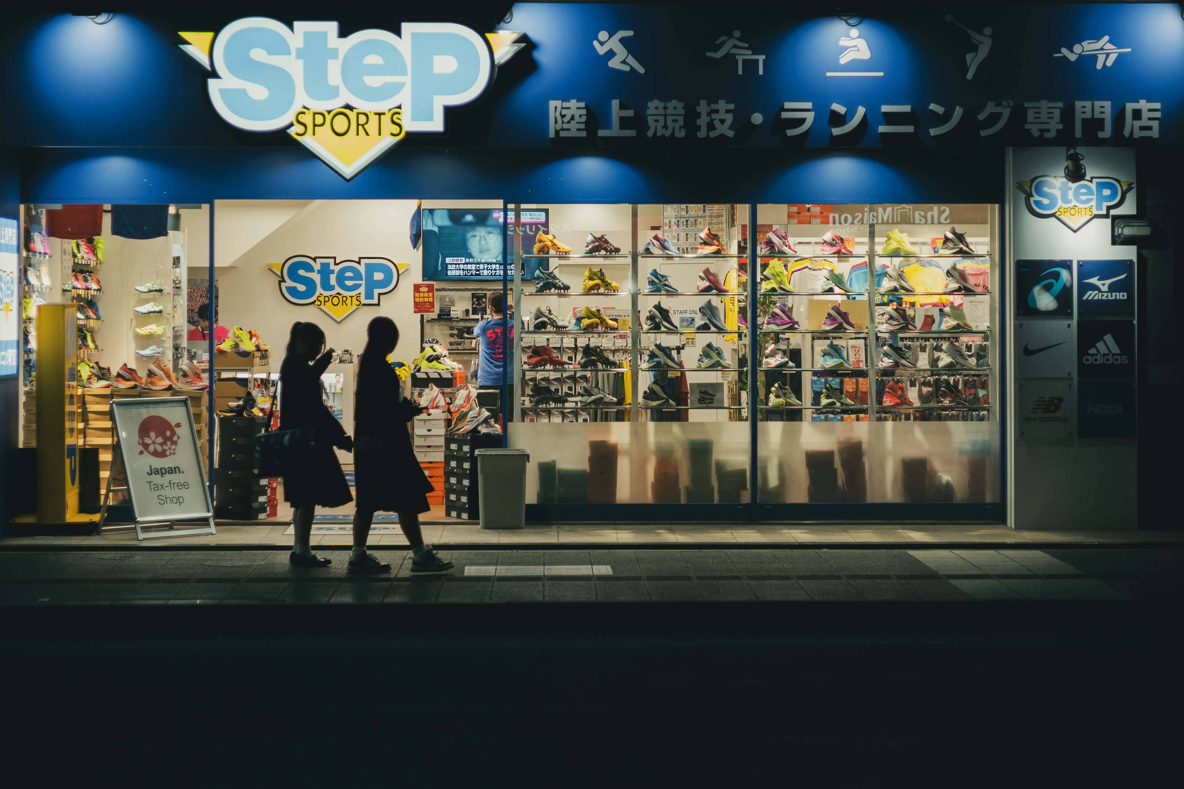 Silhouettes of two people walking in front of a brightly lit sports store at night