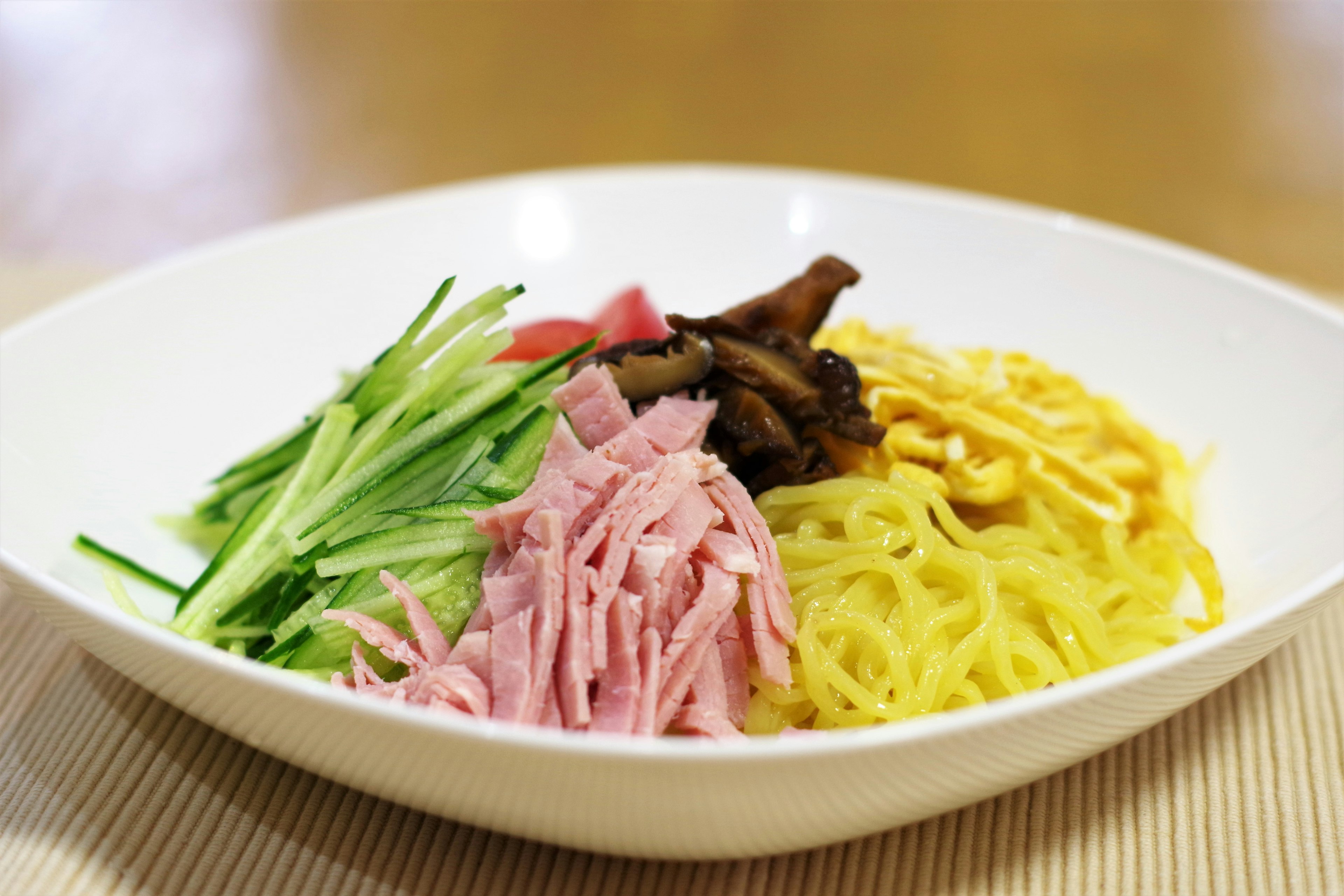 A bowl of noodles topped with sliced ham, cucumber, and yellow egg