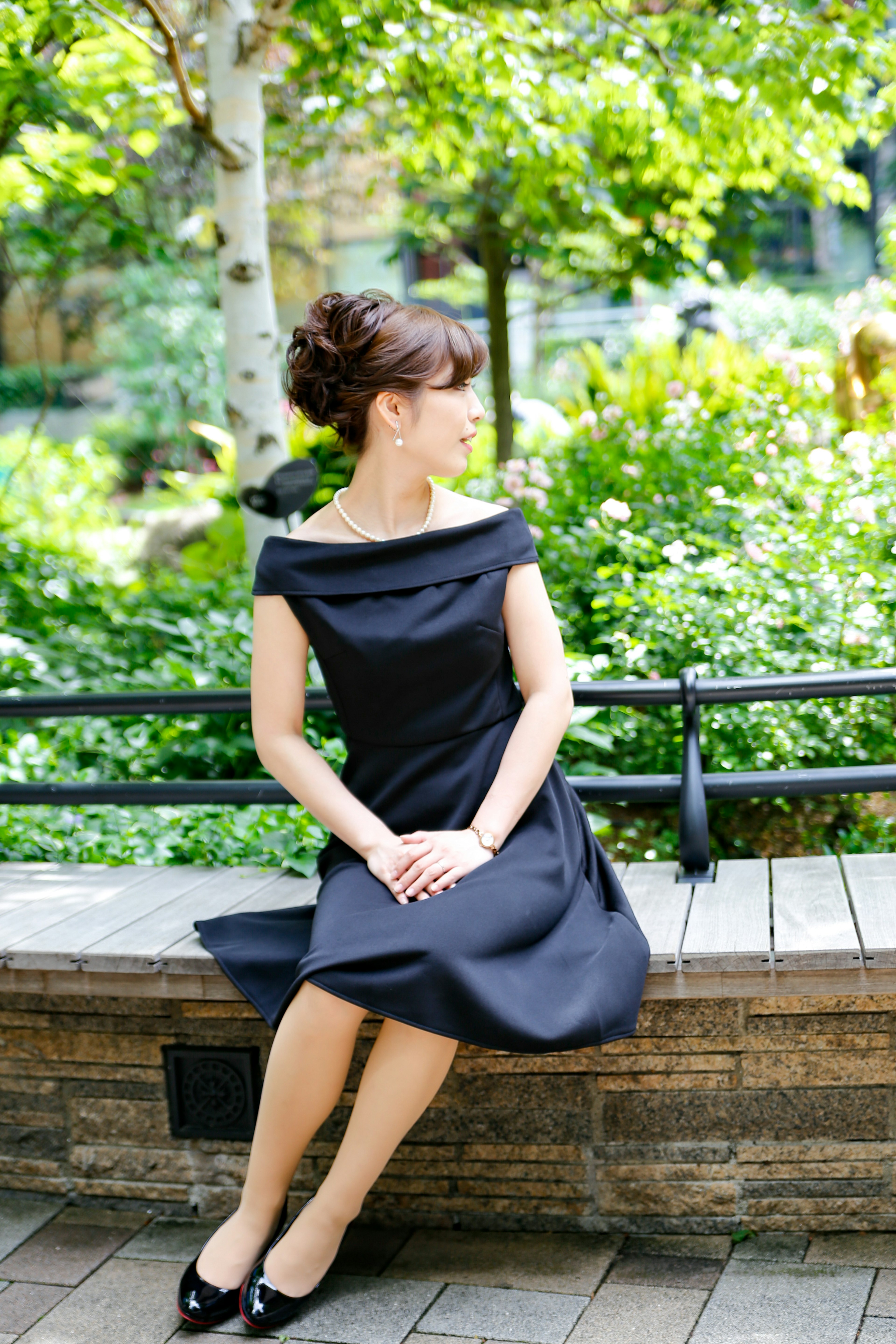 Woman sitting on a bench in a green park wearing a black dress and high heels with styled hair