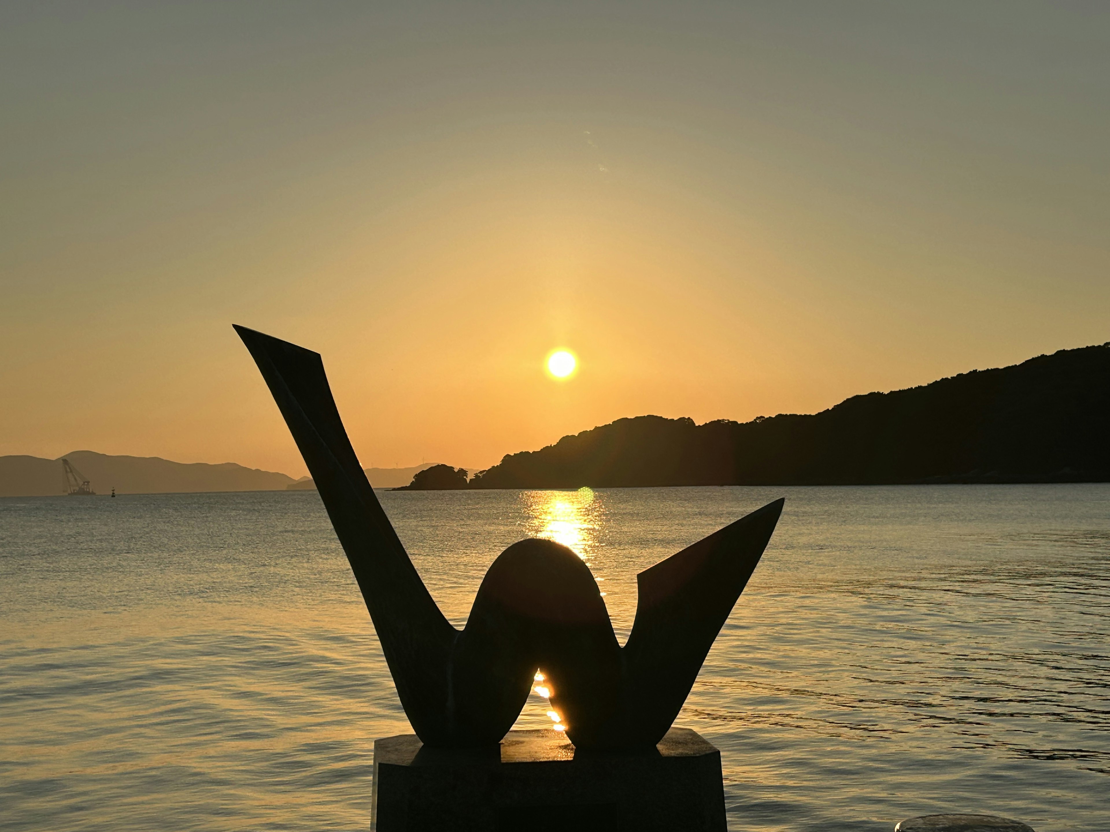 Silueta de una escultura contra un atardecer sobre el mar y las montañas