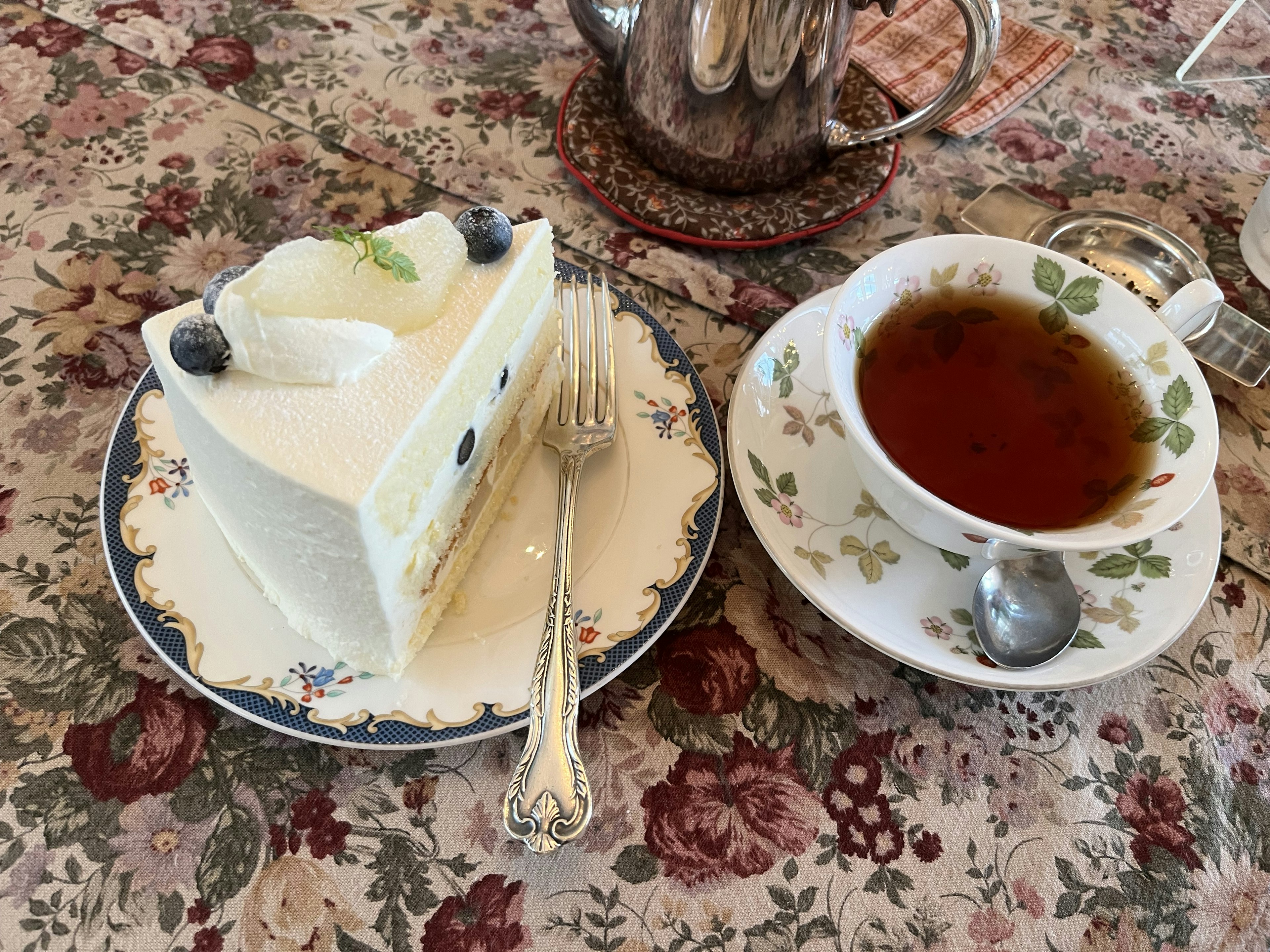 A slice of white cake with blueberries and a cup of tea on a floral tablecloth