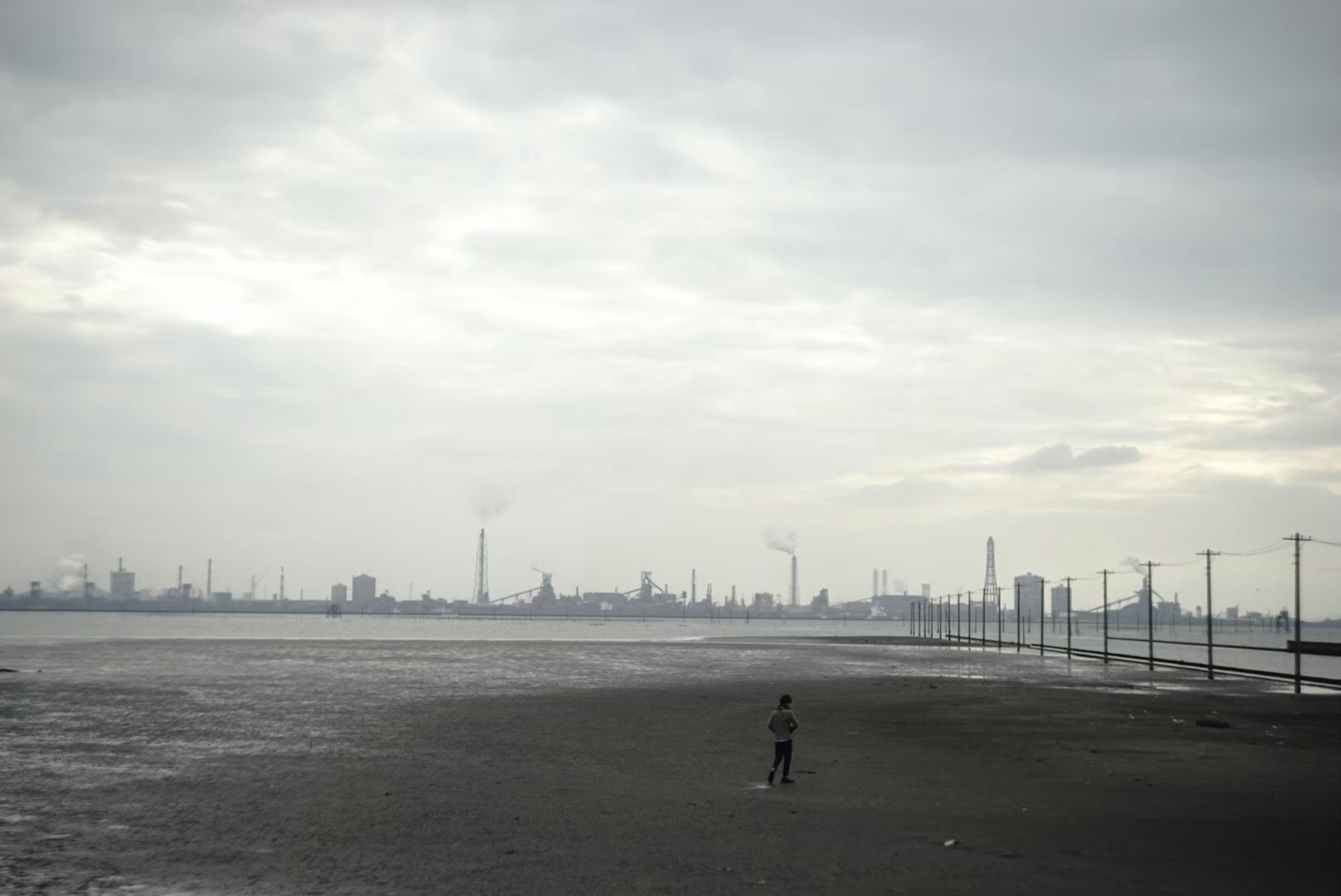 Eine einsame Figur an einem Küstenstrand mit einer Industrieansicht unter einem bewölkten Himmel