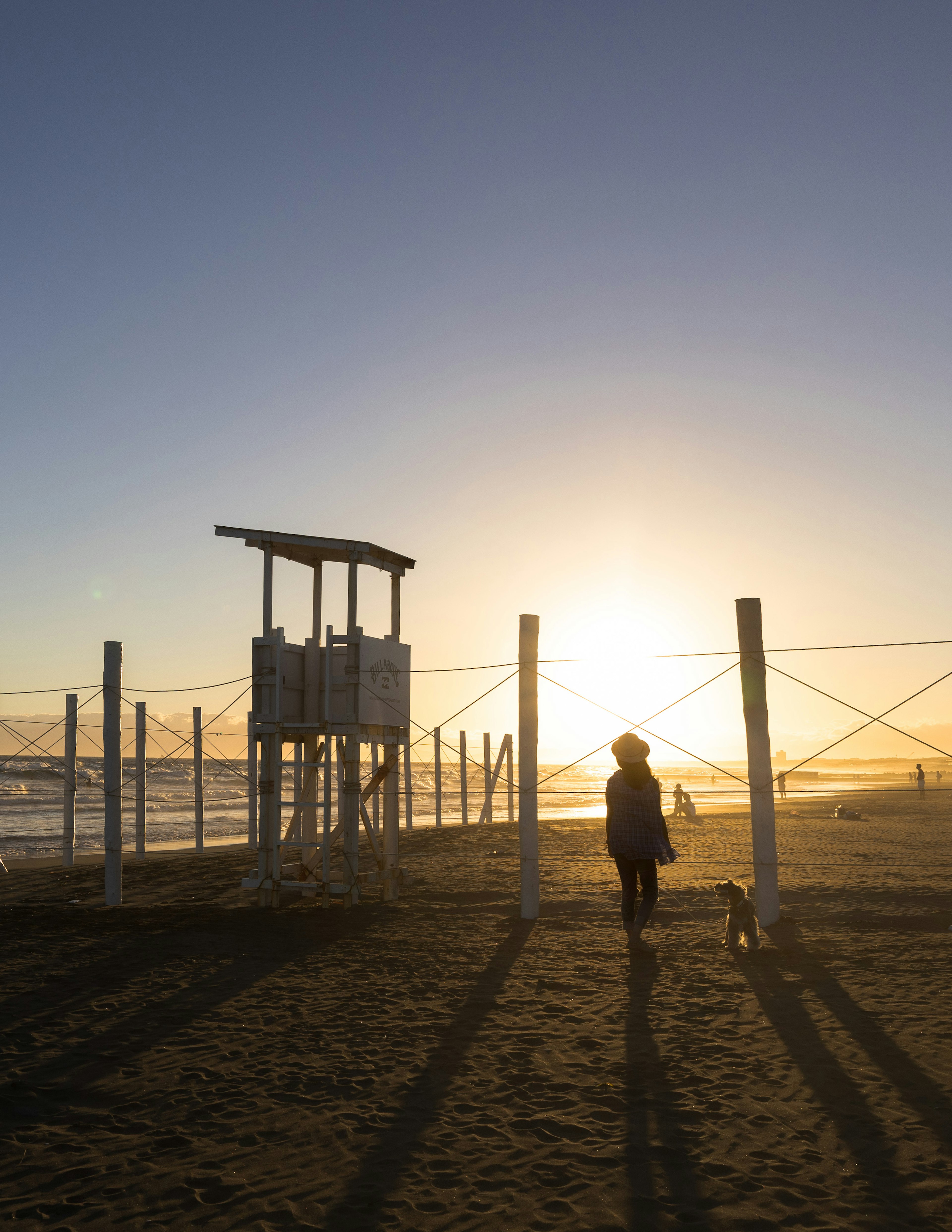 Eine Frau, die mit einem Hund am Strand bei Sonnenuntergang spaziert, mit einem Rettungsturm im Hintergrund