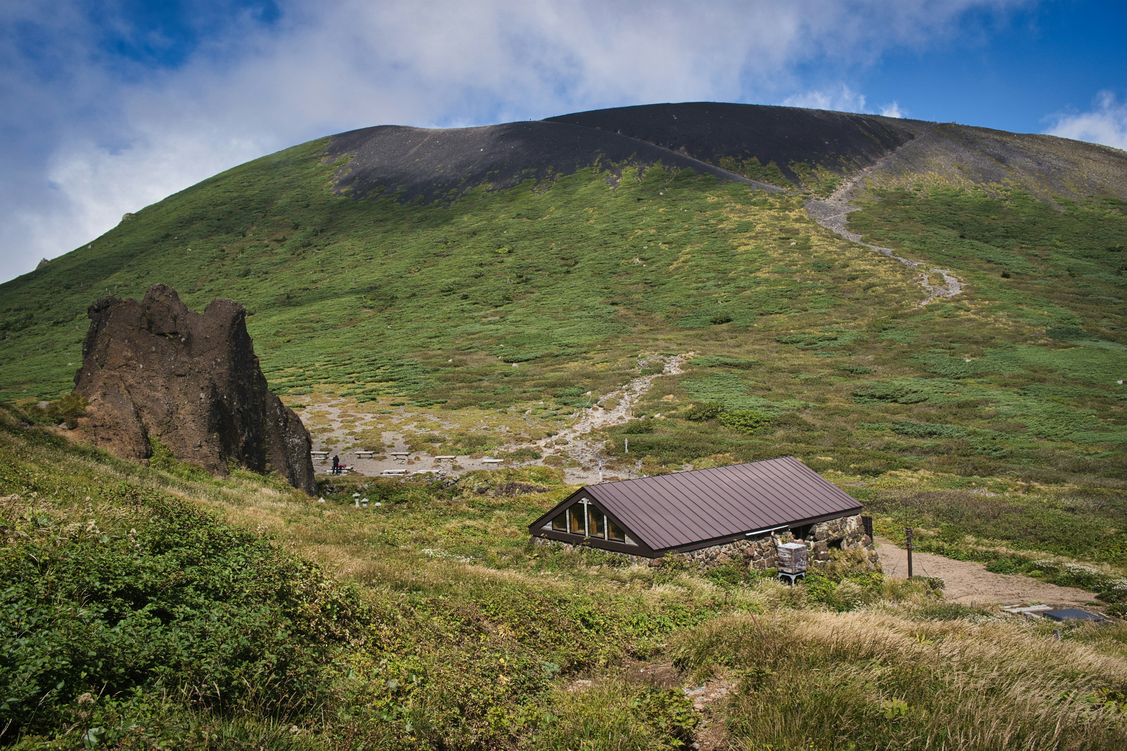 An old building at the foot of a green hill with a visible trail