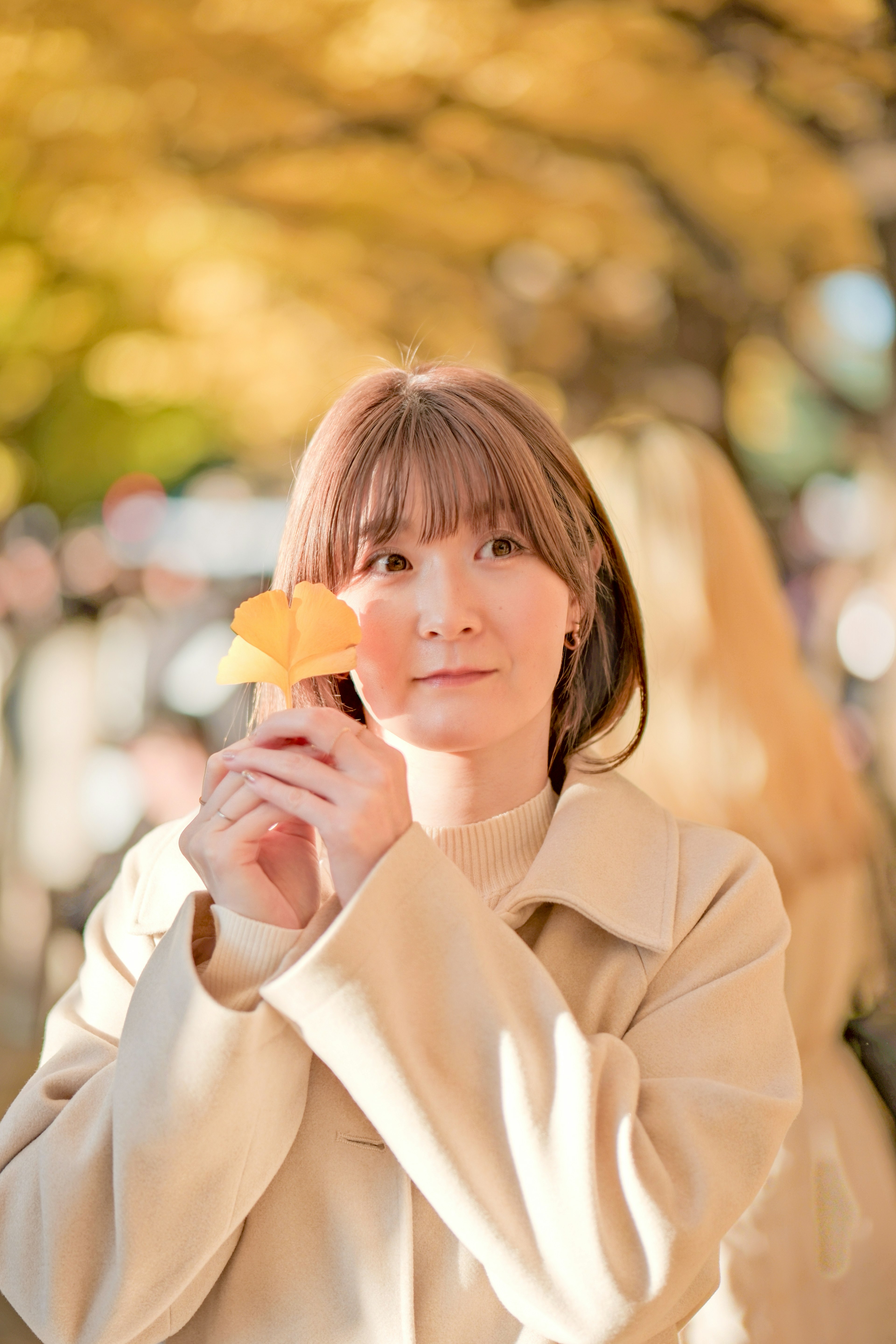 一位女性拿着一片黄色的秋天的叶子微笑，背景是银杏树