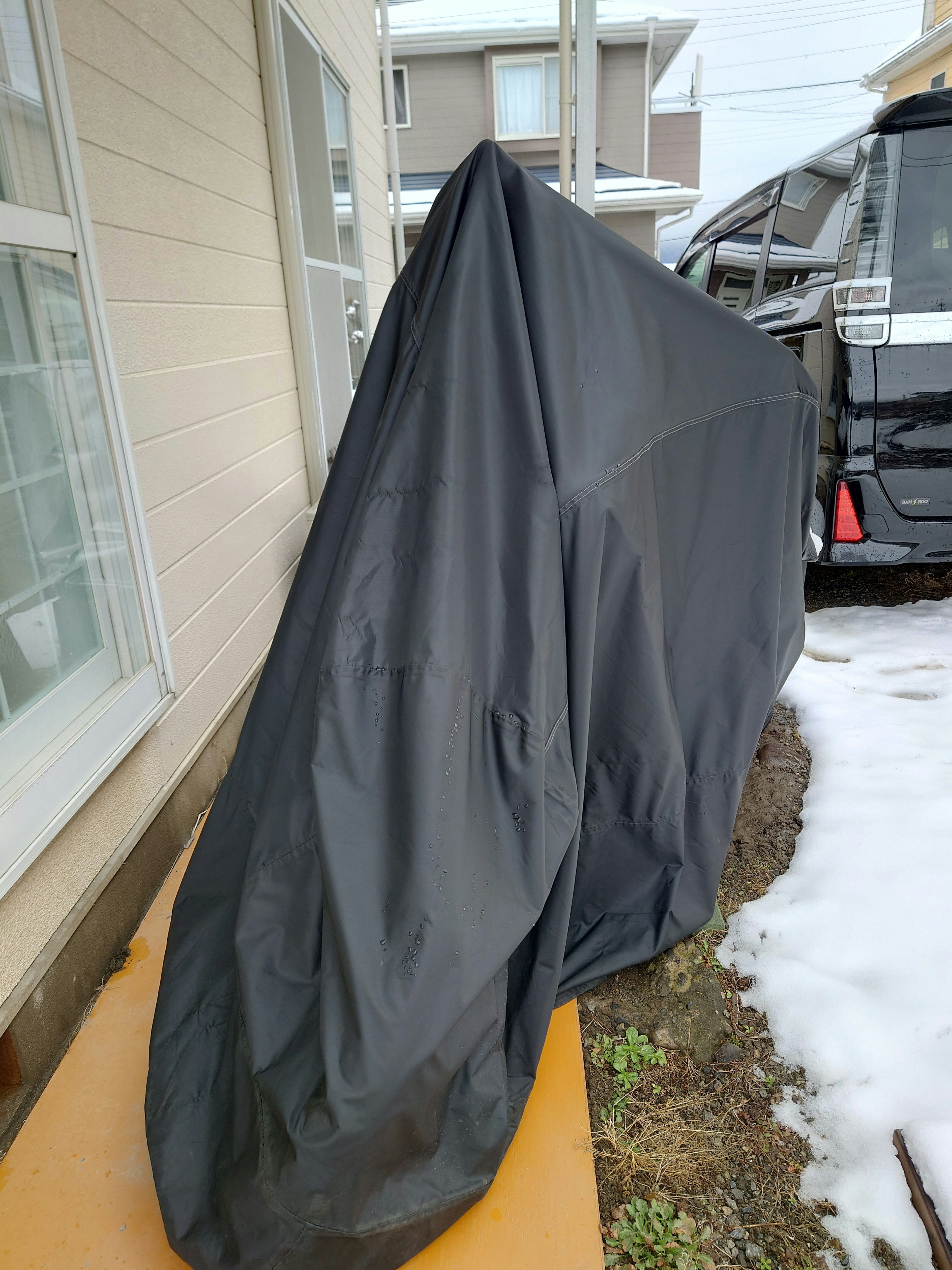 An object covered with a black tarp is outdoors with snow remaining on the ground