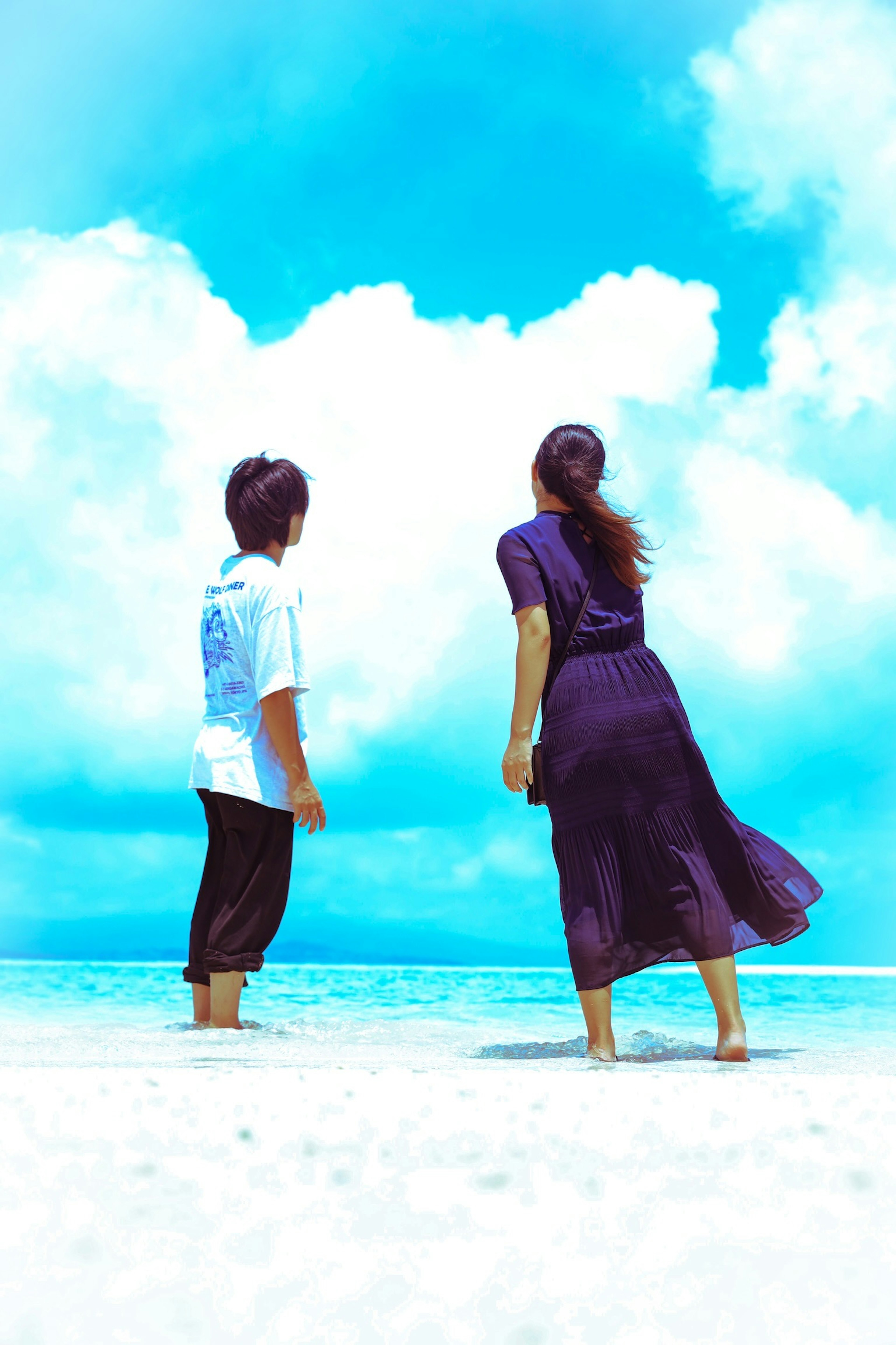 A back view of a man and woman standing by the beach with a bright blue sky and sea