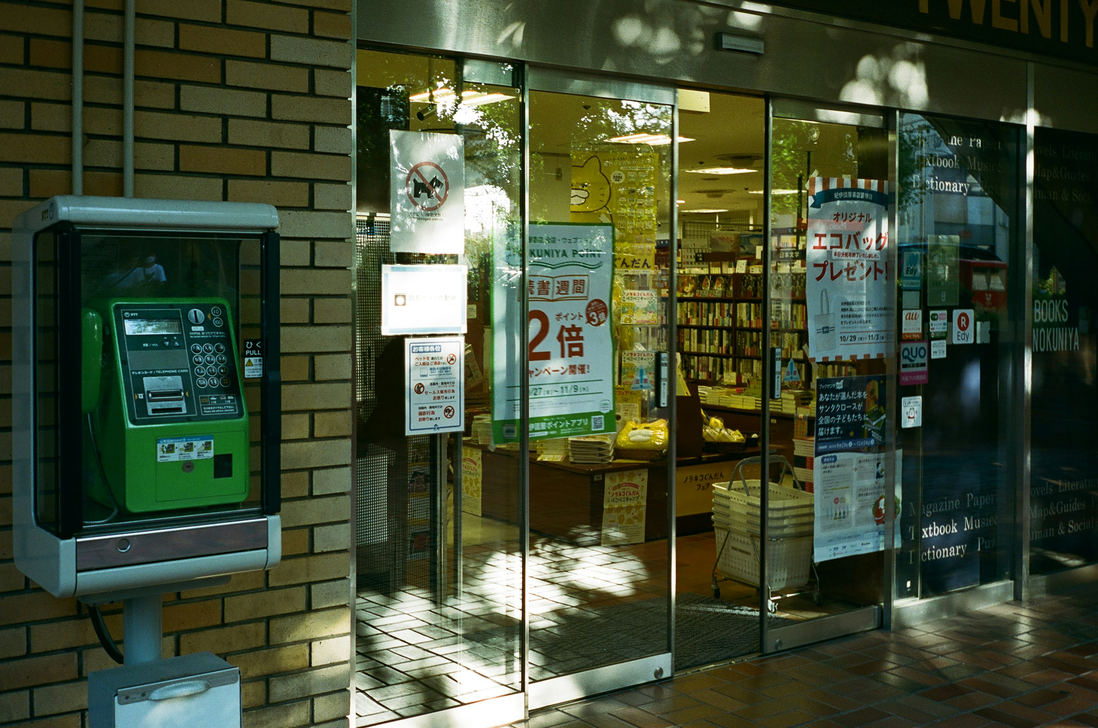 Vista esterna dell'ingresso di una biblioteca con un bancomat verde vicino a porte di vetro che mostrano scaffali di libri all'interno