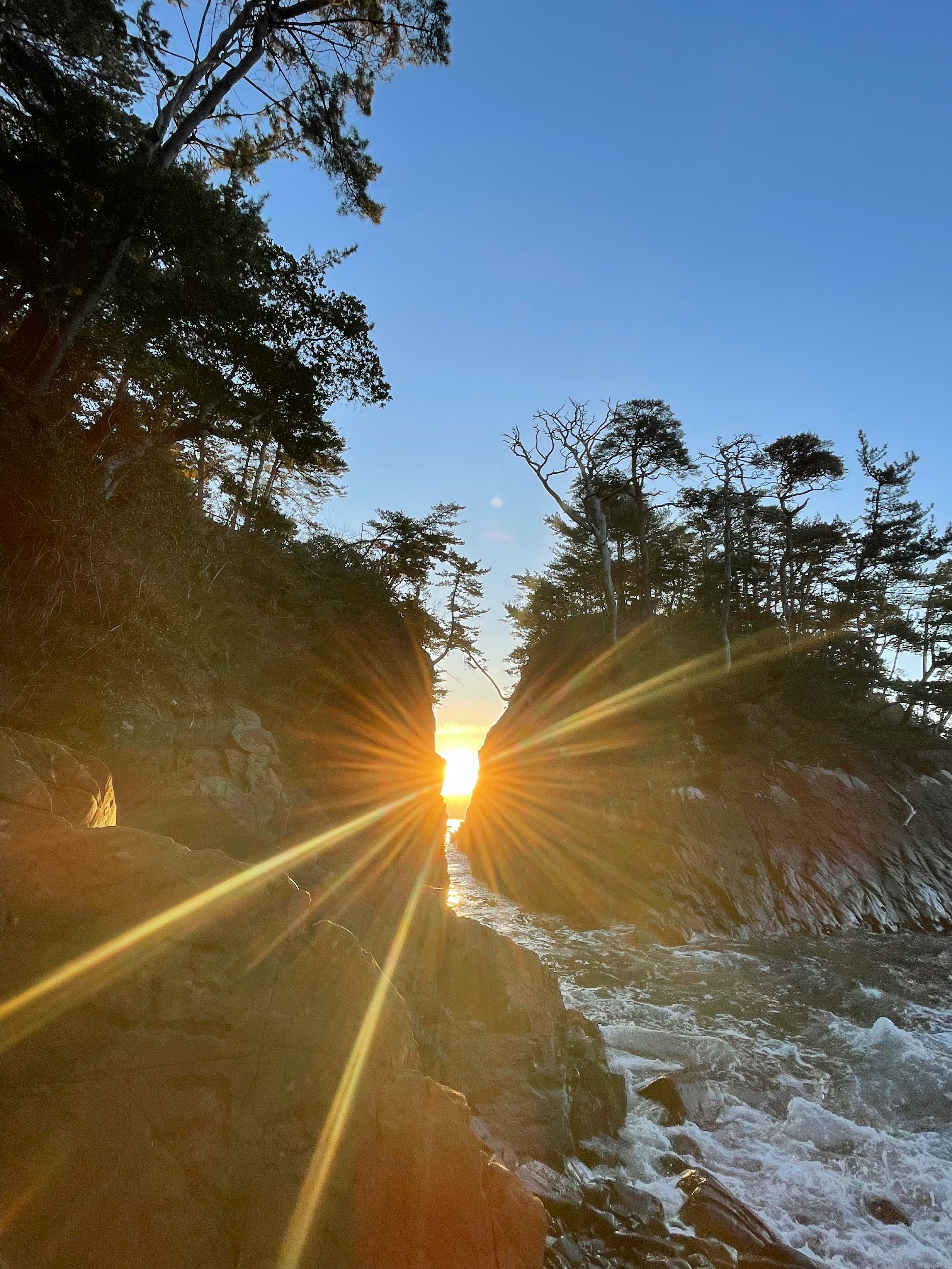 Una vista escénica que captura la puesta de sol sobre el océano enmarcada por rocas y árboles en una cala estrecha