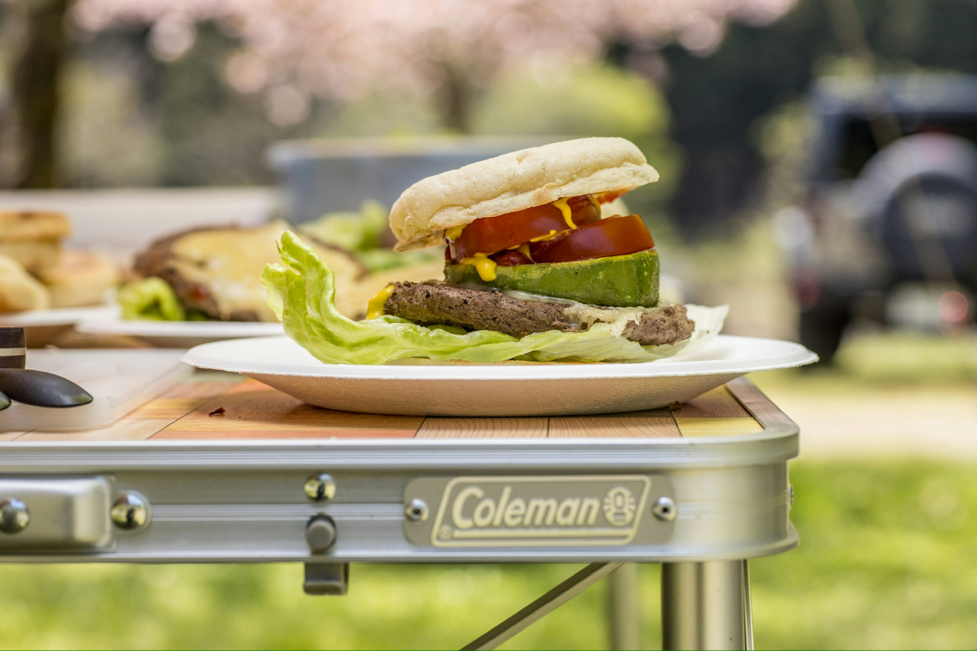 A plate with a hamburger and lettuce on a camping table with cherry blossom trees in the background