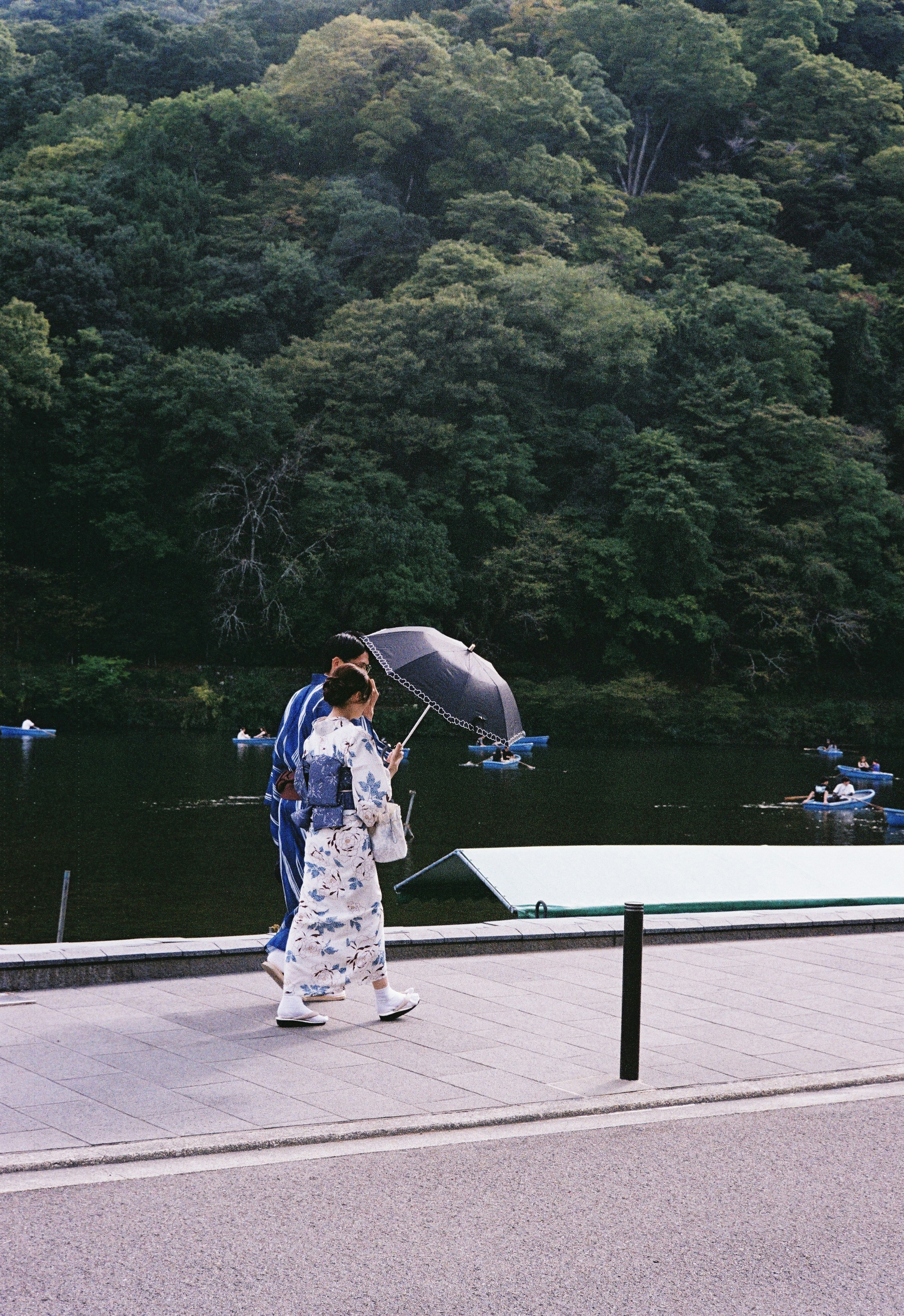 A woman in a kimono walking with an umbrella by a river surrounded by greenery