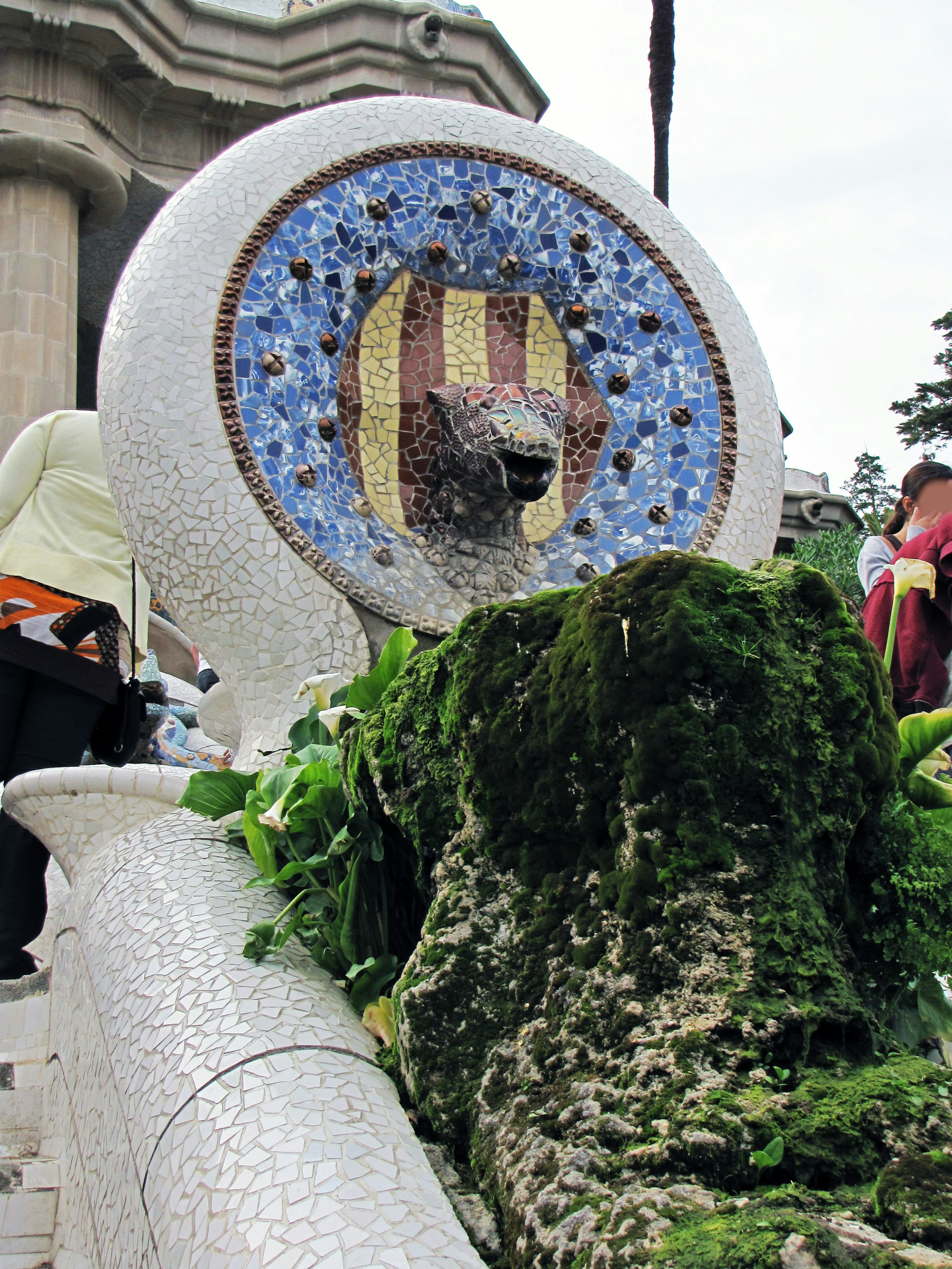 Escultura de león con ojo de mosaico rodeada de plantas verdes y personas