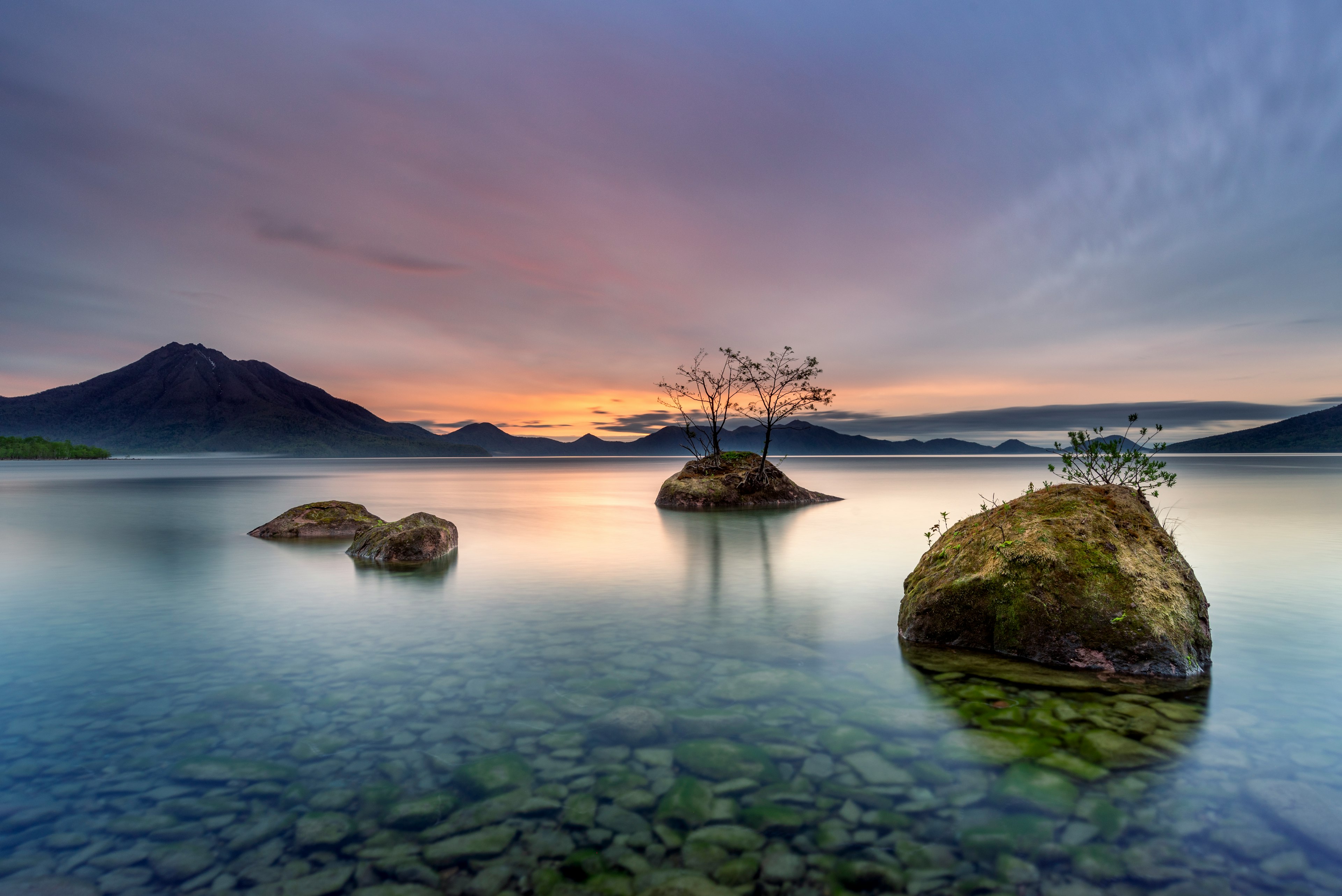 Danau tenang dengan batu dan pohon kecil mengapung di atas air, latar belakang matahari terbenam yang indah